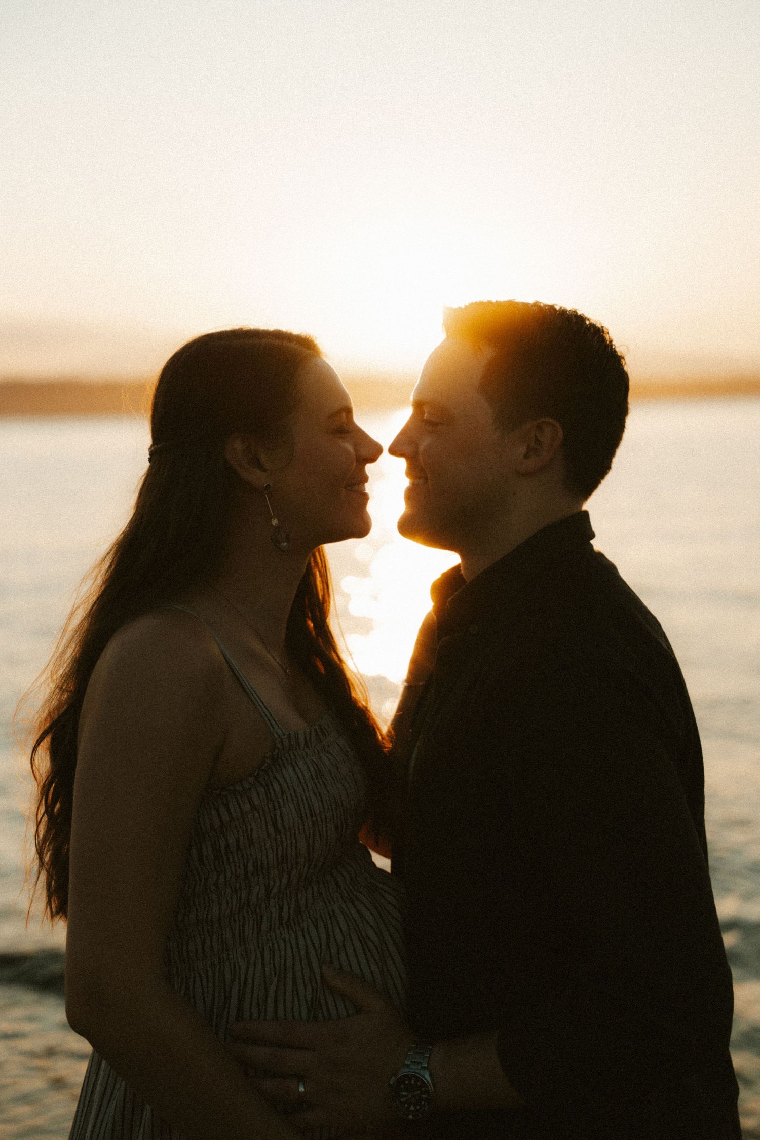 Pregnant couple sharing a romantic moment at sunset by the water, silhouetted against golden light.