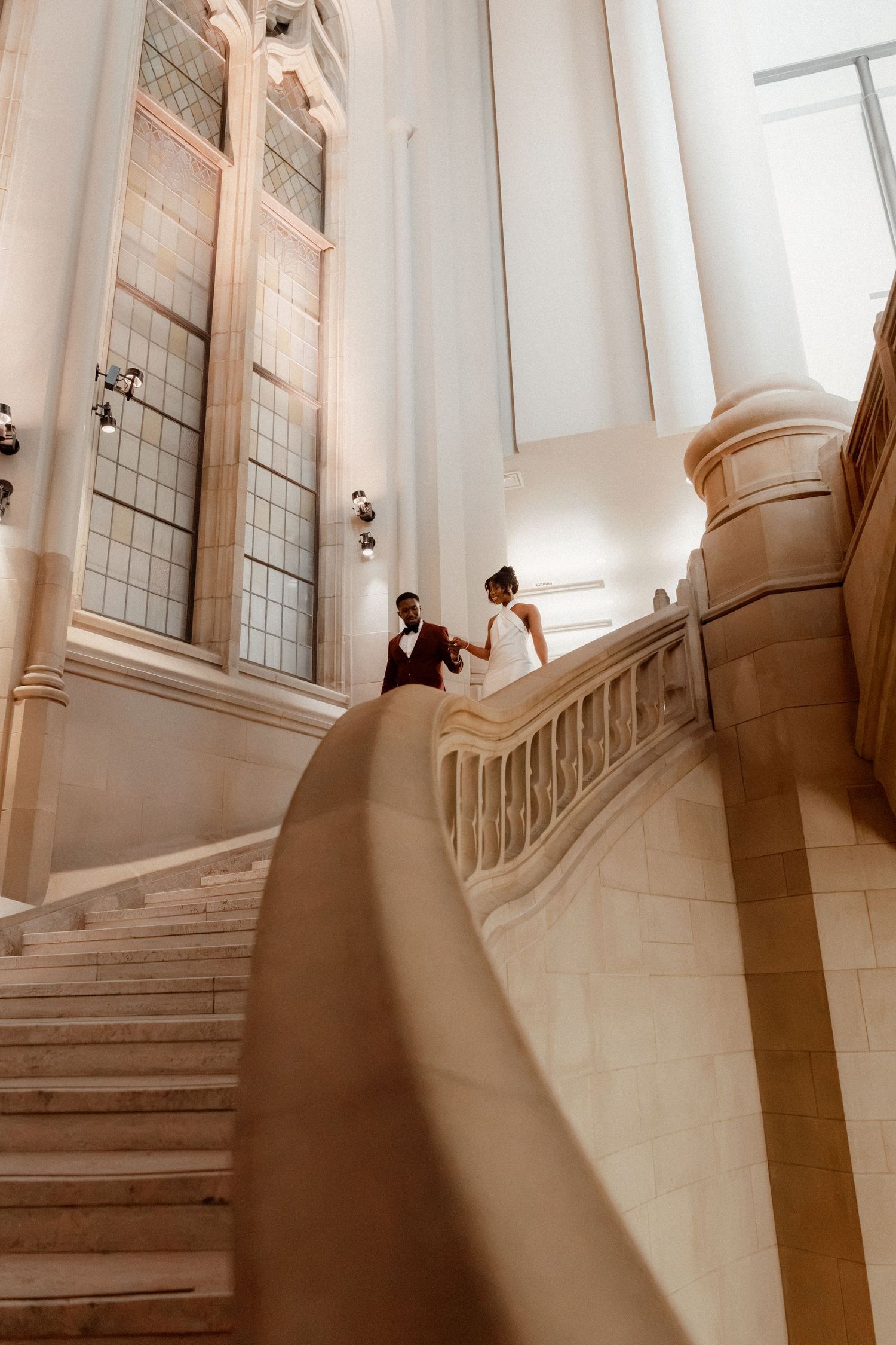 Bride and groom walking up elegant marble staircase in grand architectural venue with tall windows.