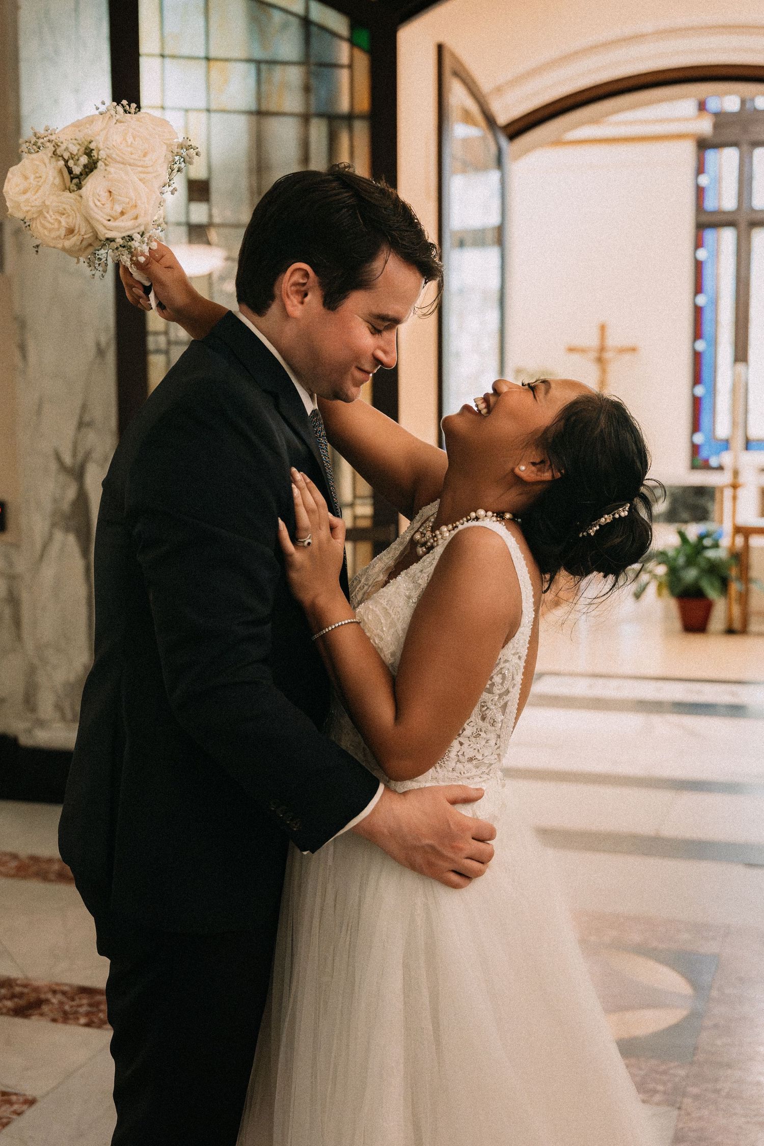 A joyful bride and groom share a romantic embrace inside a church, with the bride laughing and holding a white bouquet.