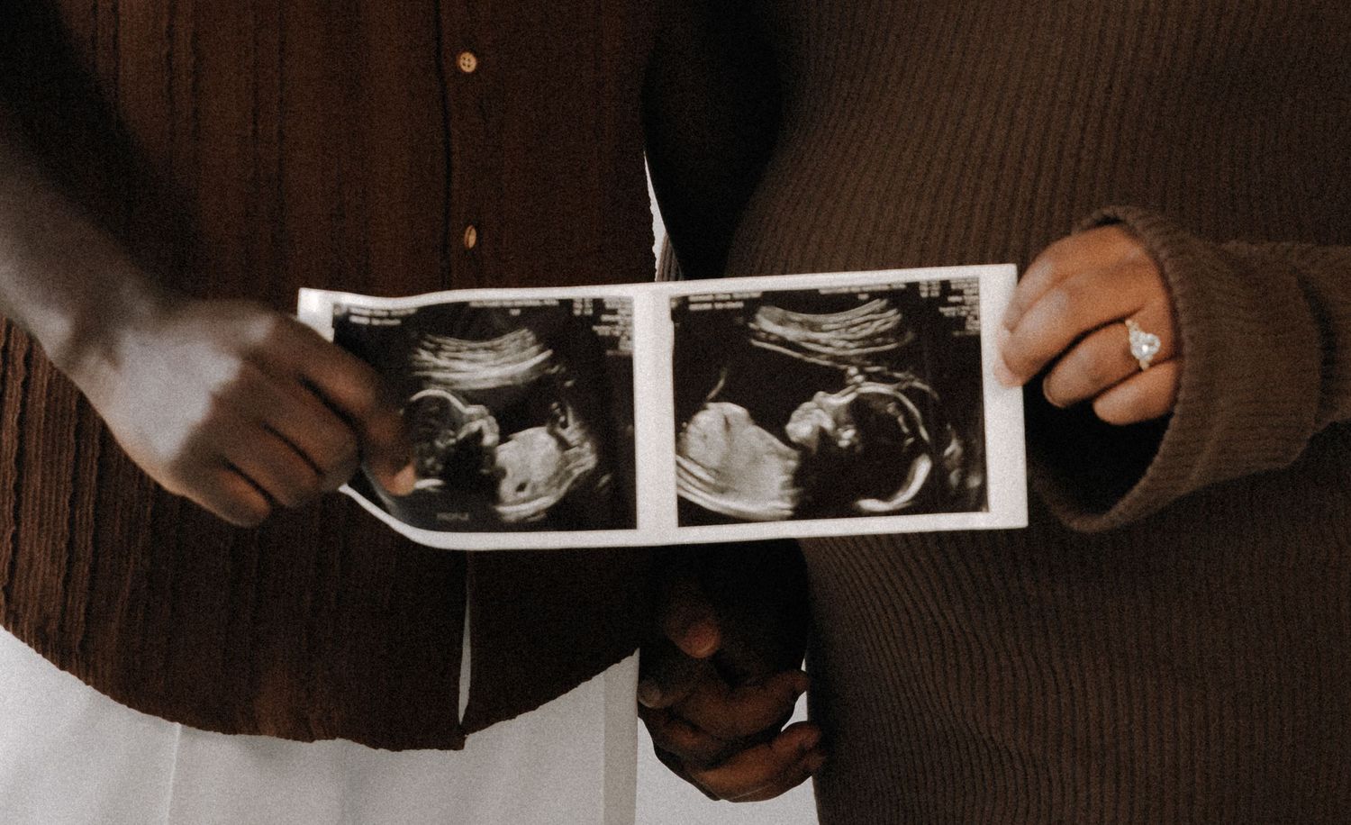Couple holding ultrasound sonogram photos showing twins, wearing brown sweaters, pregnancy announcement moment.