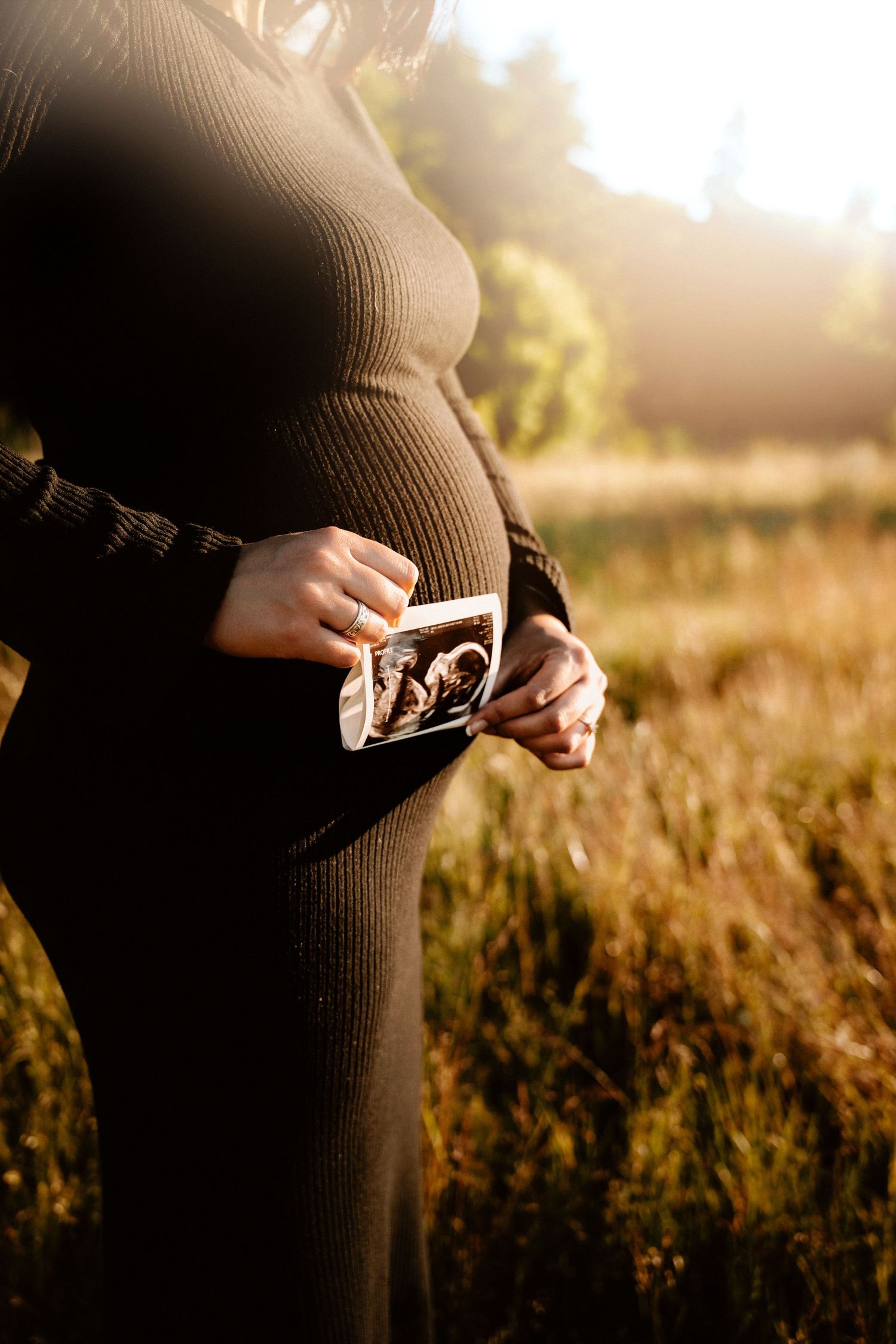 Pregnant woman in a black dress holding an ultrasound photo outdoors in golden sunlight. Seattle and Tacoma
