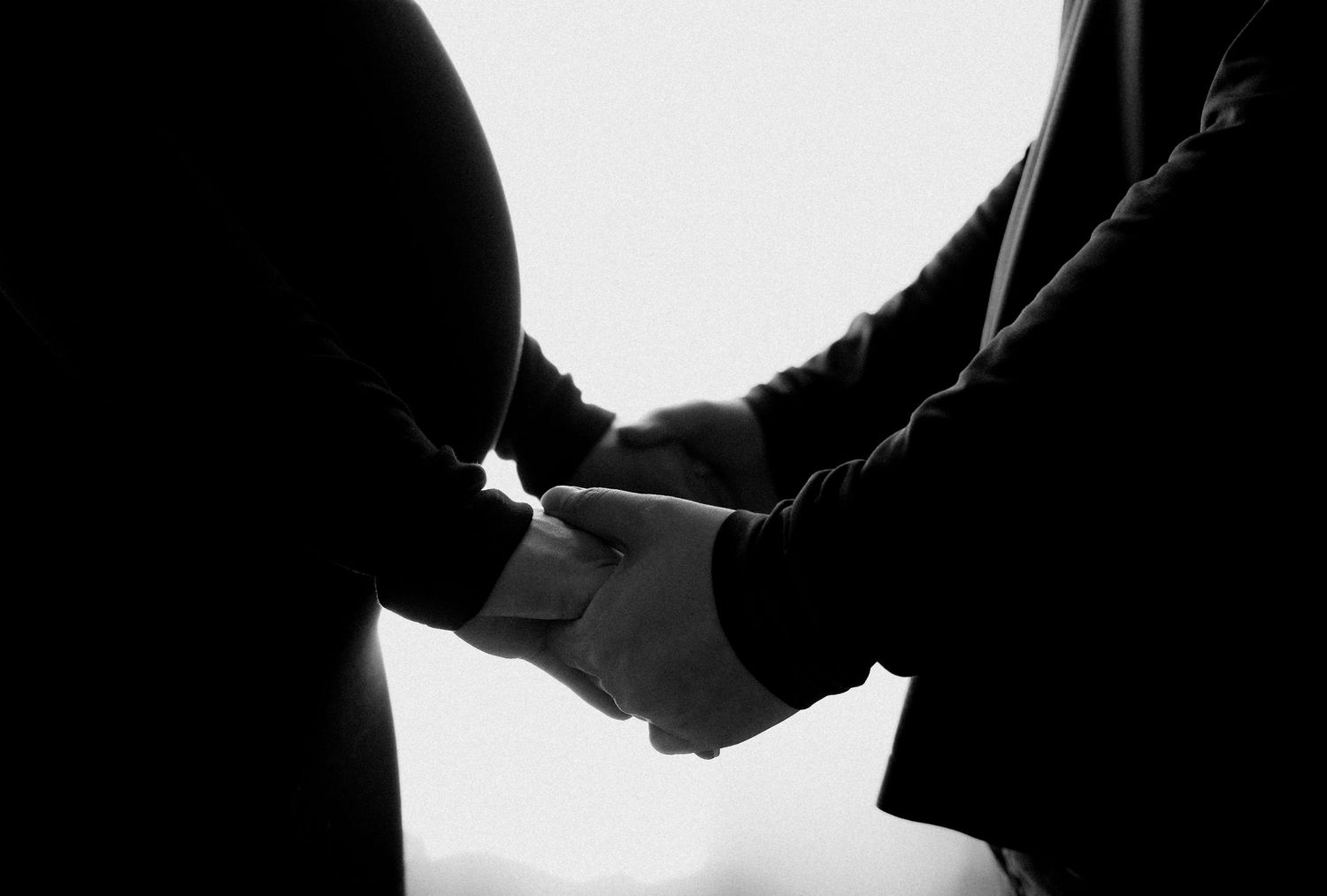 Black and white photo of pregnant woman holding hands with partner, silhouette maternity portrait.