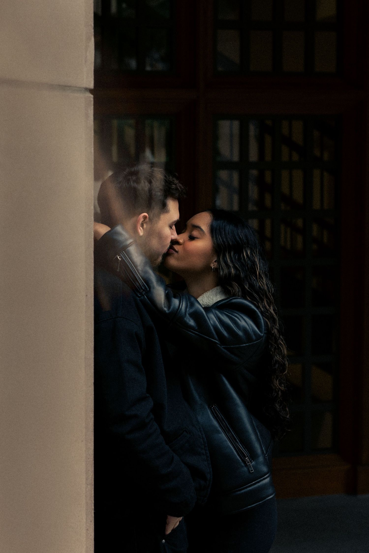 Couple sharing a romantic kiss leaning against a stone pillar in moody urban setting.