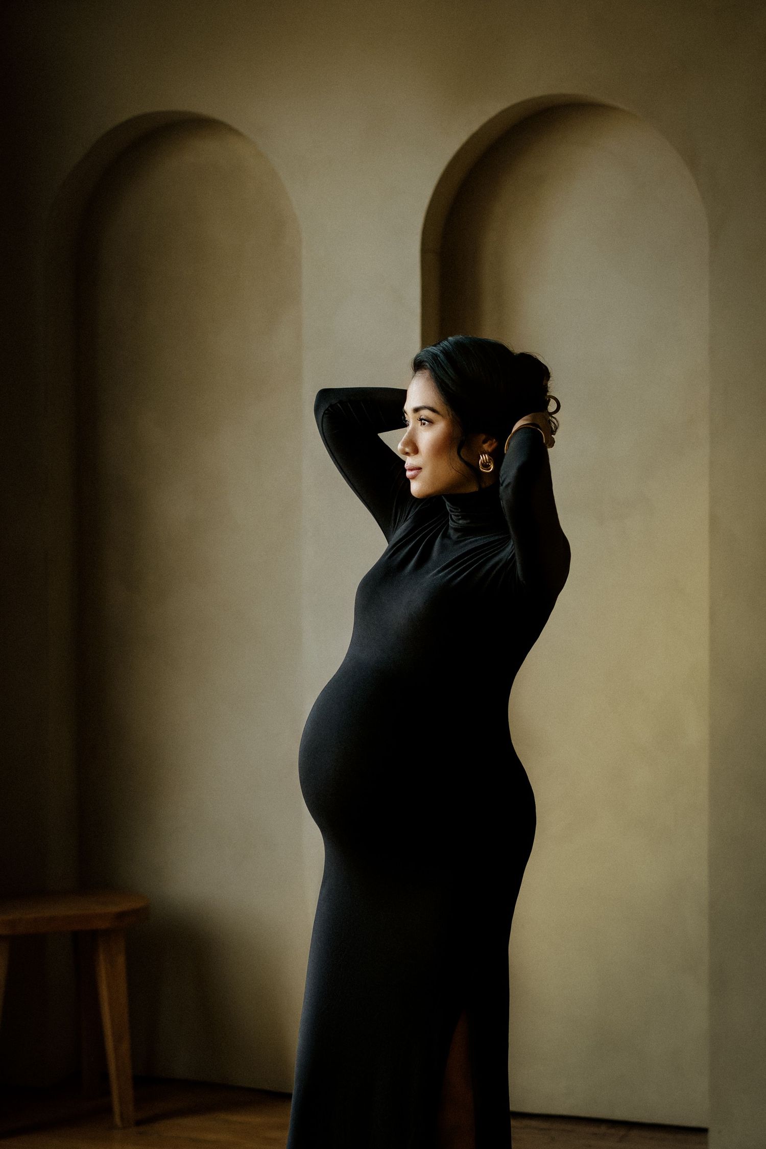 Elegant pregnant woman in a black turtleneck dress posing gracefully in front of arched wall niches.