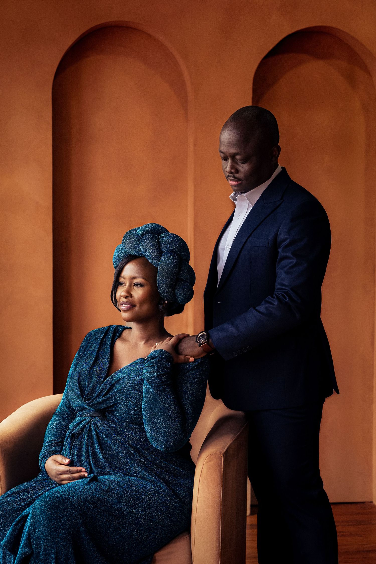Elegant pregnant woman in teal dress and headwrap sits while man in navy suit stands beside her.