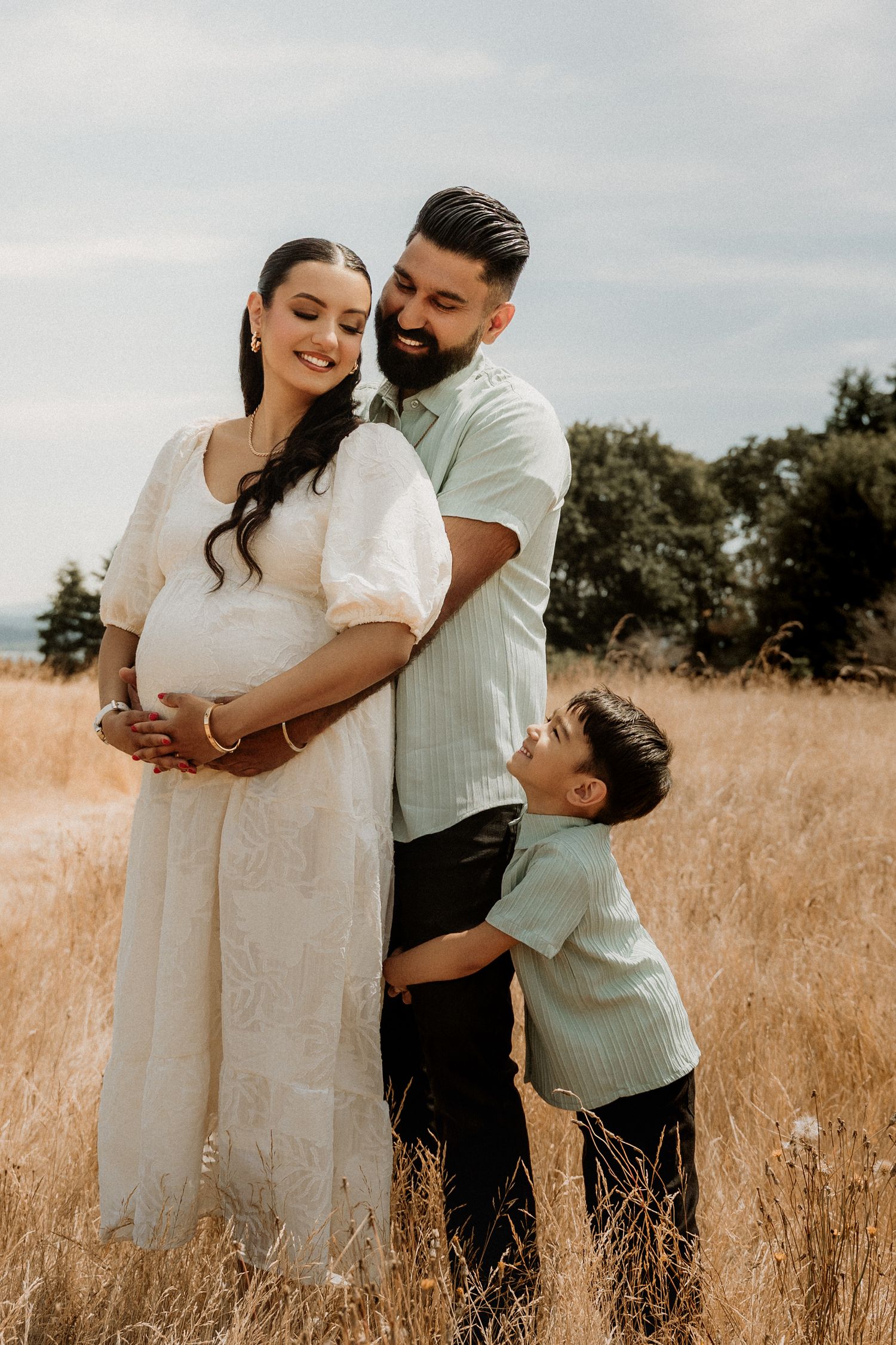 Pregnant woman in white dress with husband and young son posing in golden field for maternity photo shoot.