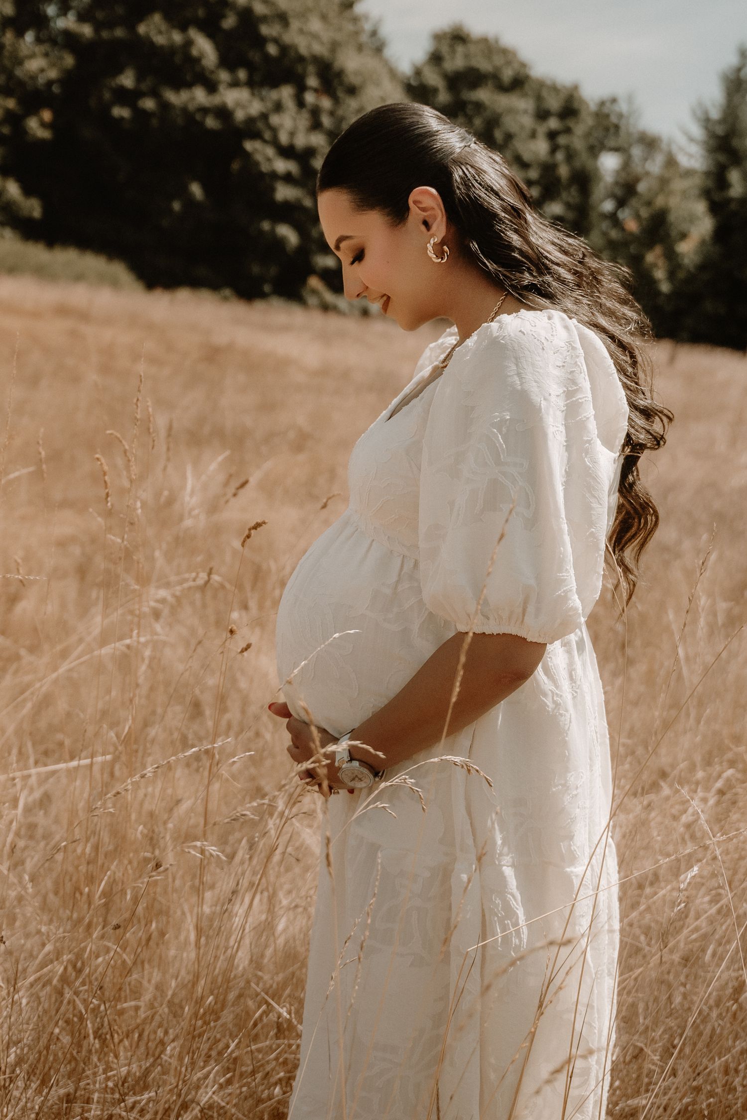 Pregnant woman in white dress standing in golden field, cradling baby bump, with flowing dark hair and trees behind.