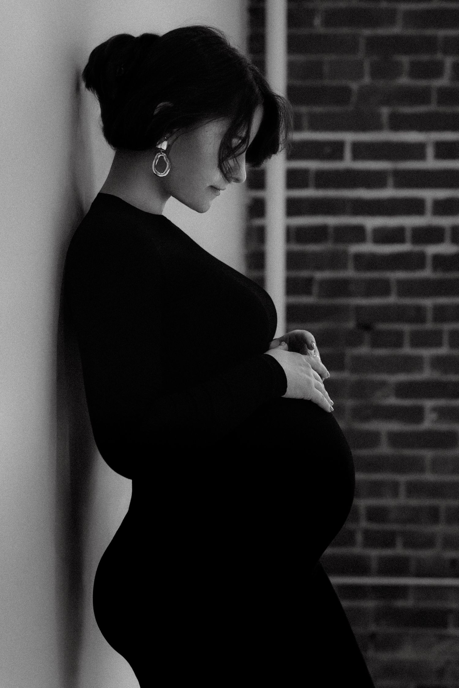 Black and white profile portrait of a pregnant woman in a black dress, cradling her baby bump against a brick wall.
