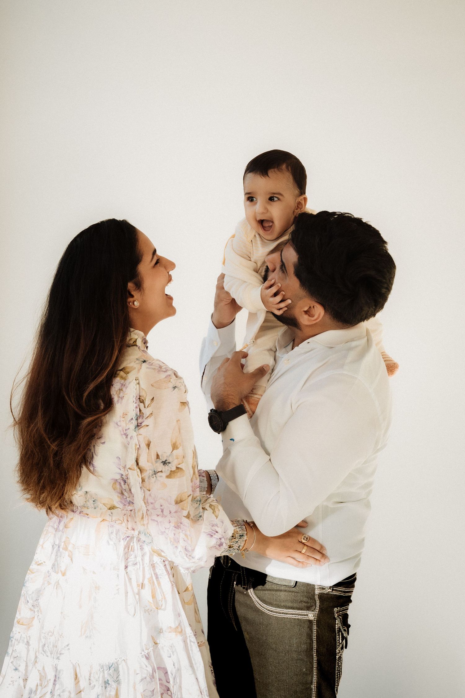 Happy family portrait with mother in floral dress, father in white shirt, and baby in bright studio setting.