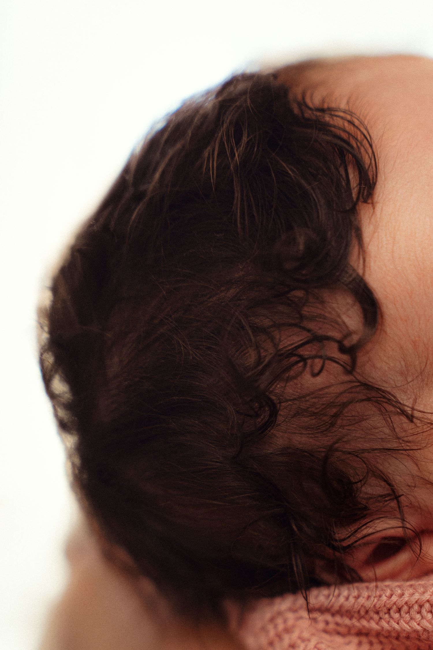 Close-up of a newborn baby with dark curly hair wearing a pink outfit against a white background.