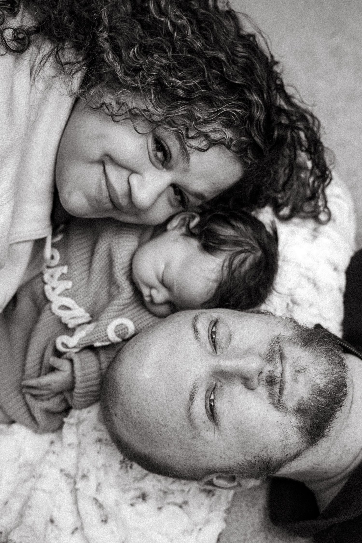 Black and white portrait of smiling curly-haired mother, newborn baby, and father lying together in tender family moment.