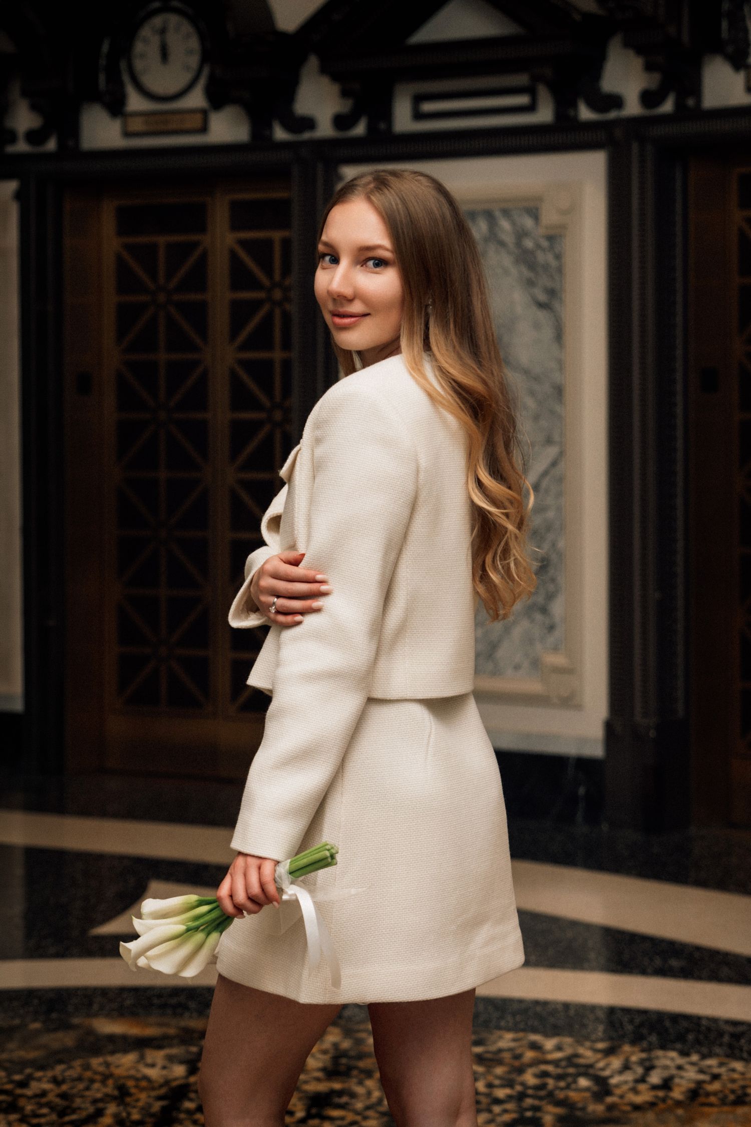 Young woman in cream white outfit holding white tulips, smiling over shoulder in elegant marble interior.