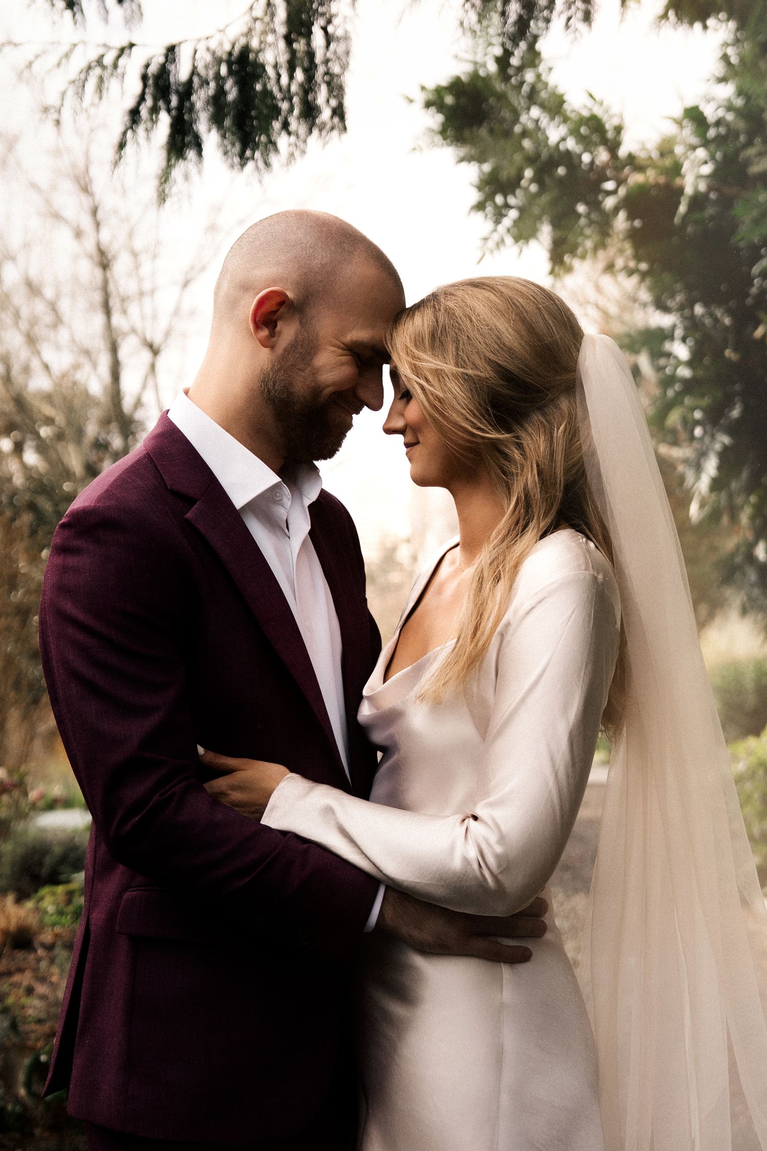 Bride and groom in intimate embrace, she wears white gown with veil, he wears burgundy suit, outdoors.
