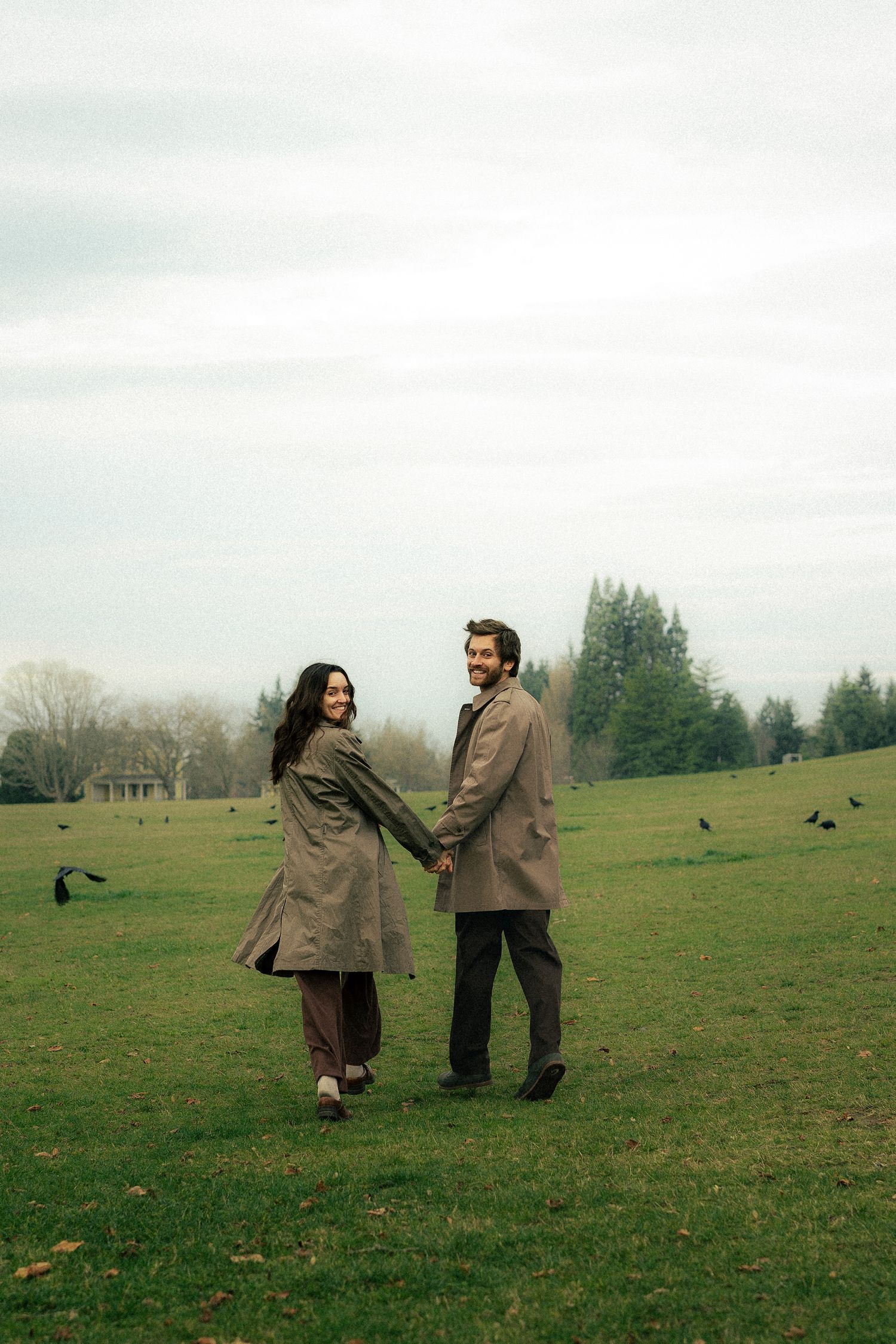 Couple holding hands walking in green park wearing matching trench coats on overcast day with birds on grass.
