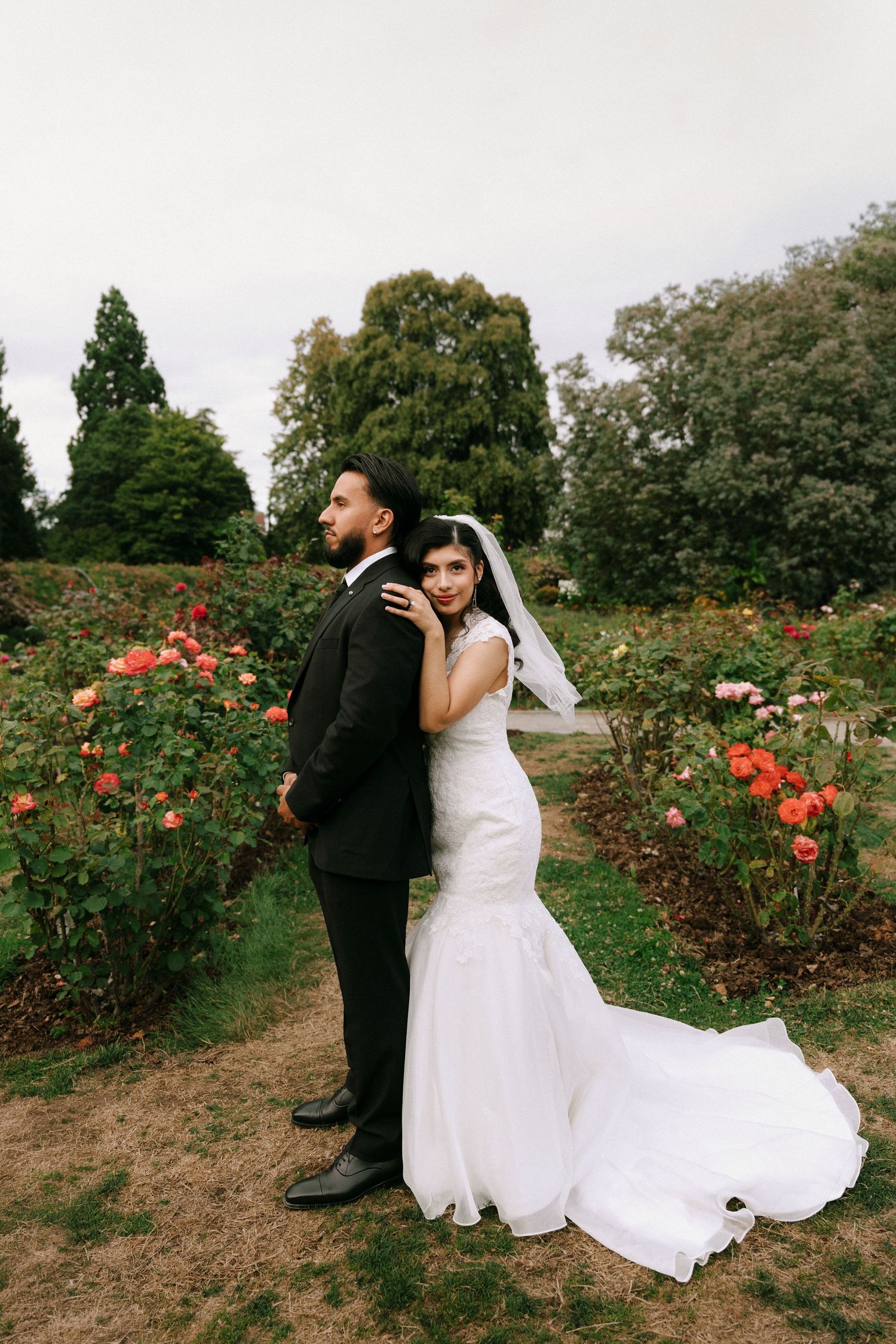 Bride and groom pose back-to-back in a rose garden, bride in mermaid gown with veil, groom in black suit.