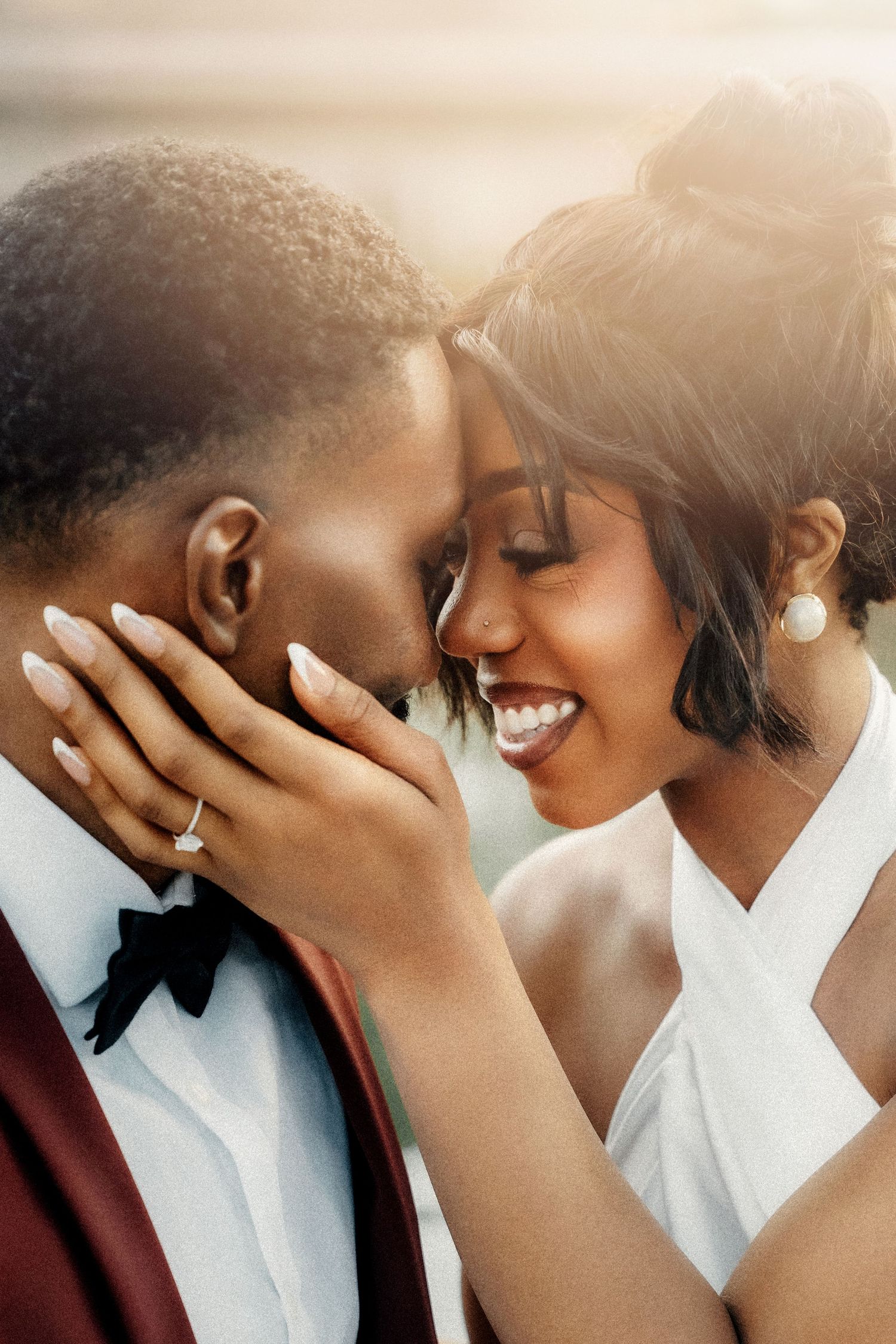 A joyful Black couple in wedding attire share a romantic forehead touch, the bride smiling in white dress with engagement ring visible.