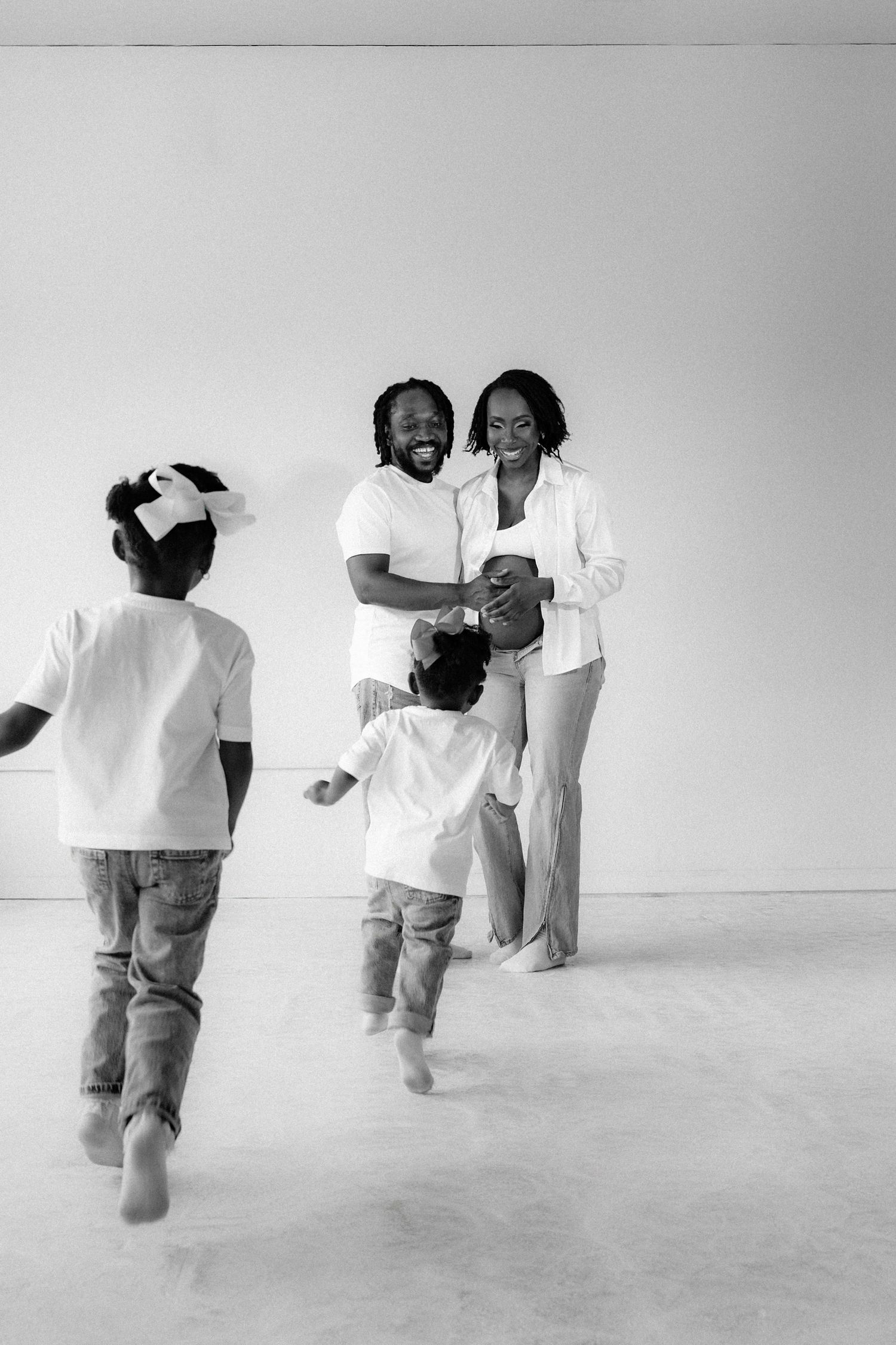 Black and white photo of a joyful family with two children running toward their smiling parents in a bright room.