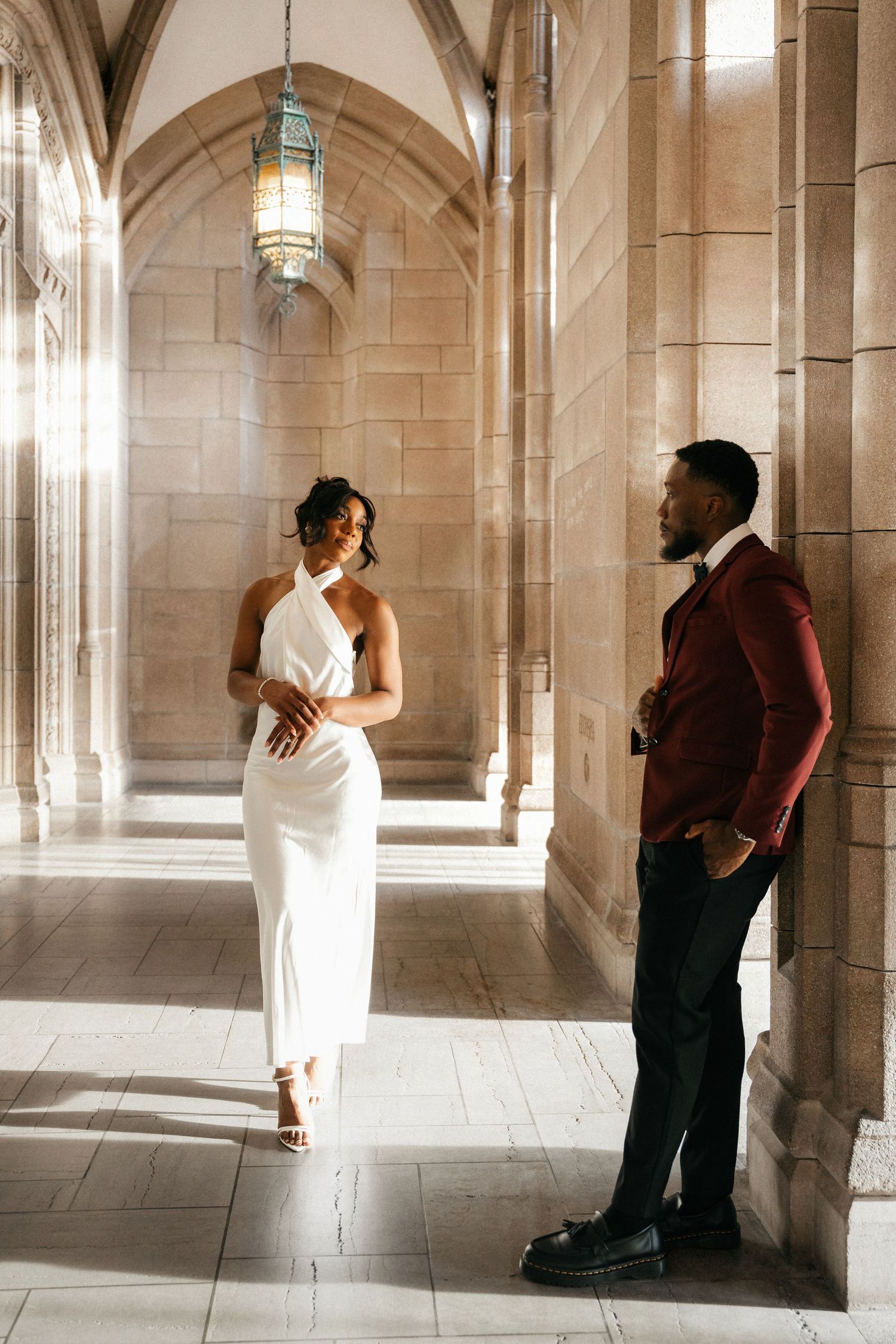 Elegant couple in a gothic stone hallway, woman in white halter dress, man in burgundy suit, sharing a romantic moment.