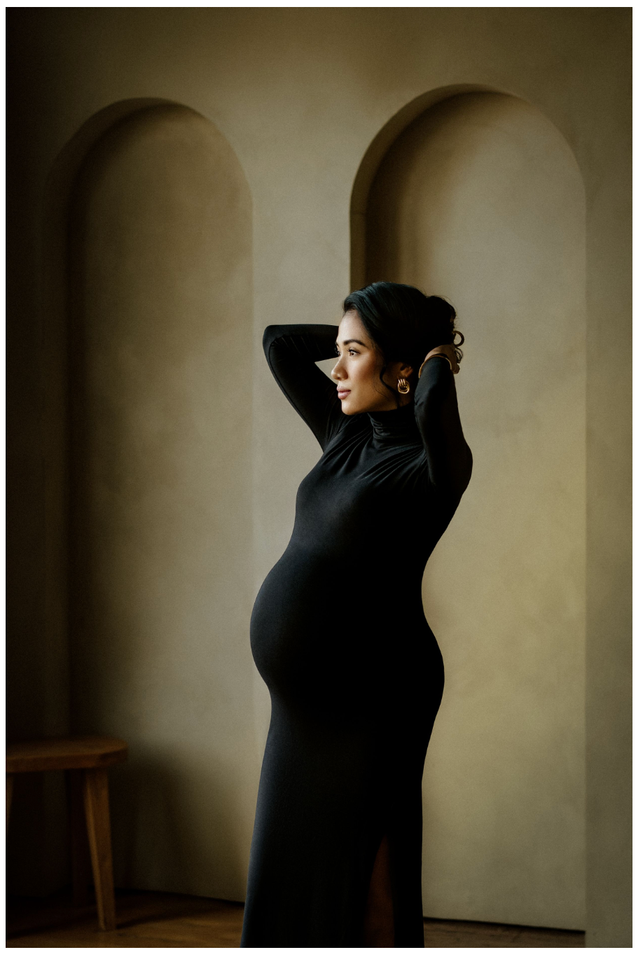 A pregnant woman in an elegant black turtleneck maxi dress poses sideways near arched wall niches in a warm studio. Seattle and Tacoma