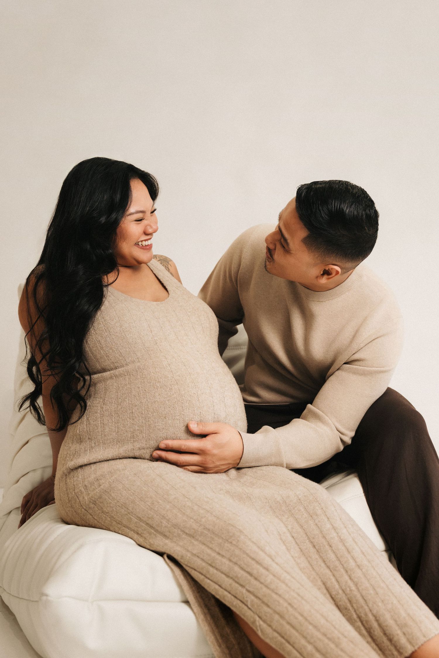 A smiling pregnant woman in a beige dress sits with her partner, both cradling her baby bump during a maternity photoshoot.