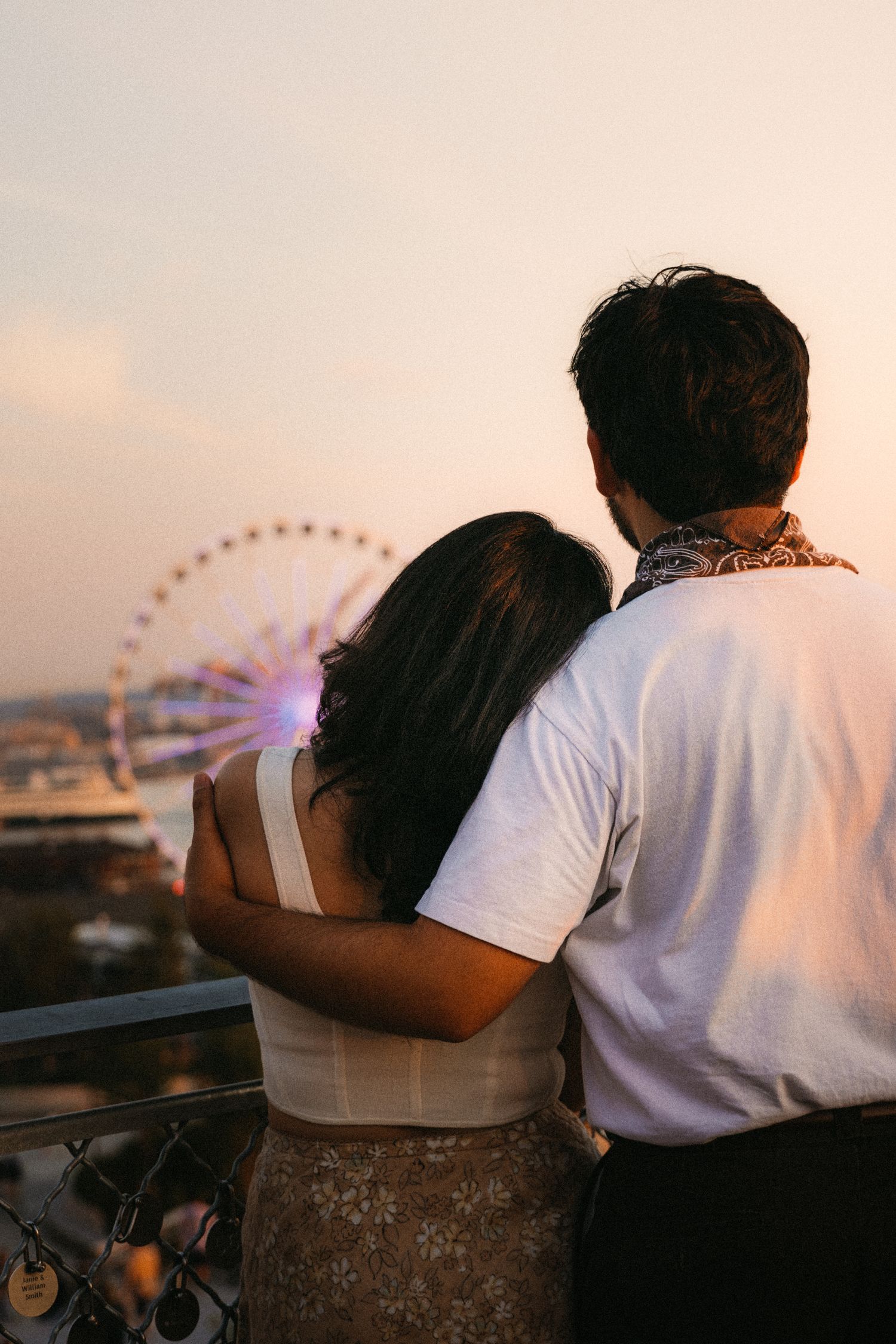 Couple embracing on a rooftop balcony at sunset, gazing at a illuminated Ferris wheel in the distance.
