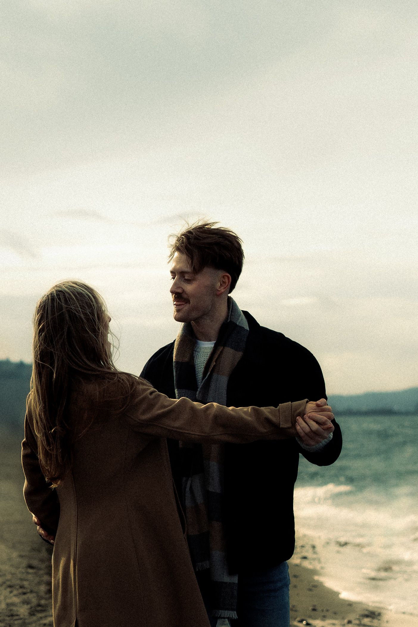 Couple dancing romantically on a beach, man smiling at woman, both wearing coats on a cloudy day.