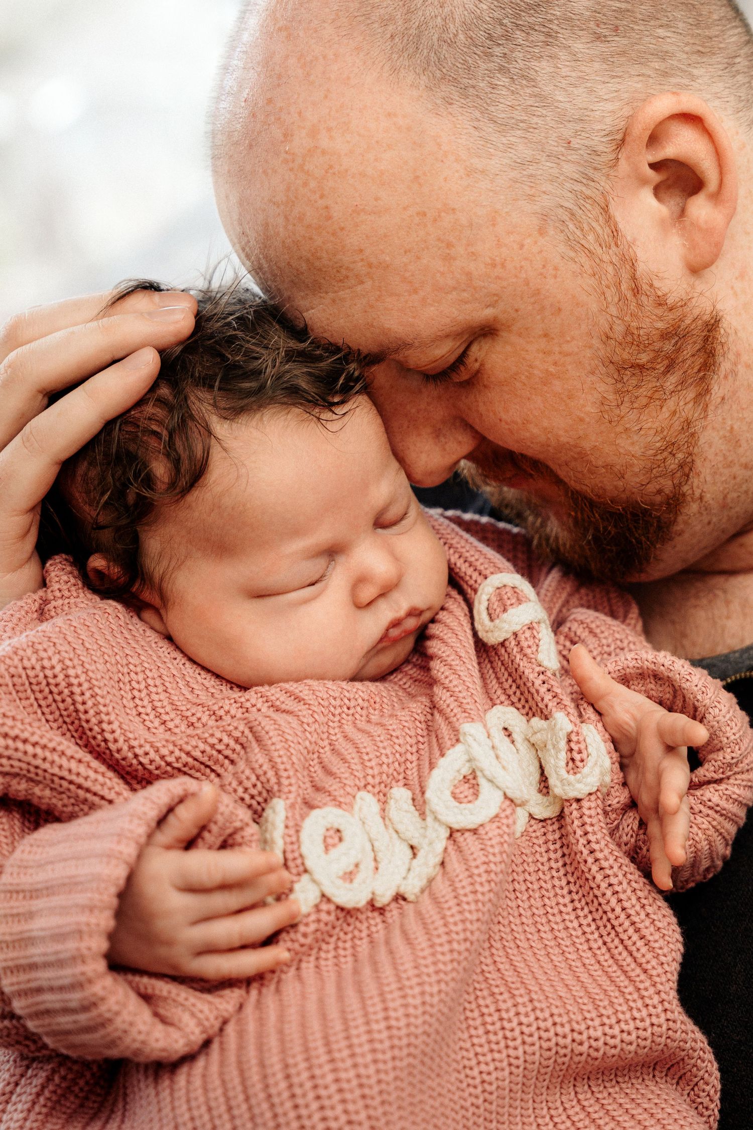 A bearded father tenderly kisses his sleeping newborn baby dressed in a pink knit sweater.