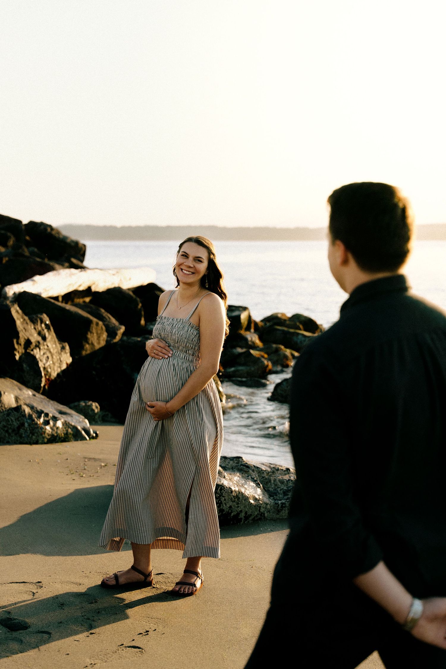 Smiling pregnant woman in striped dress holds belly on sandy beach at sunset with rocky shore backdrop.