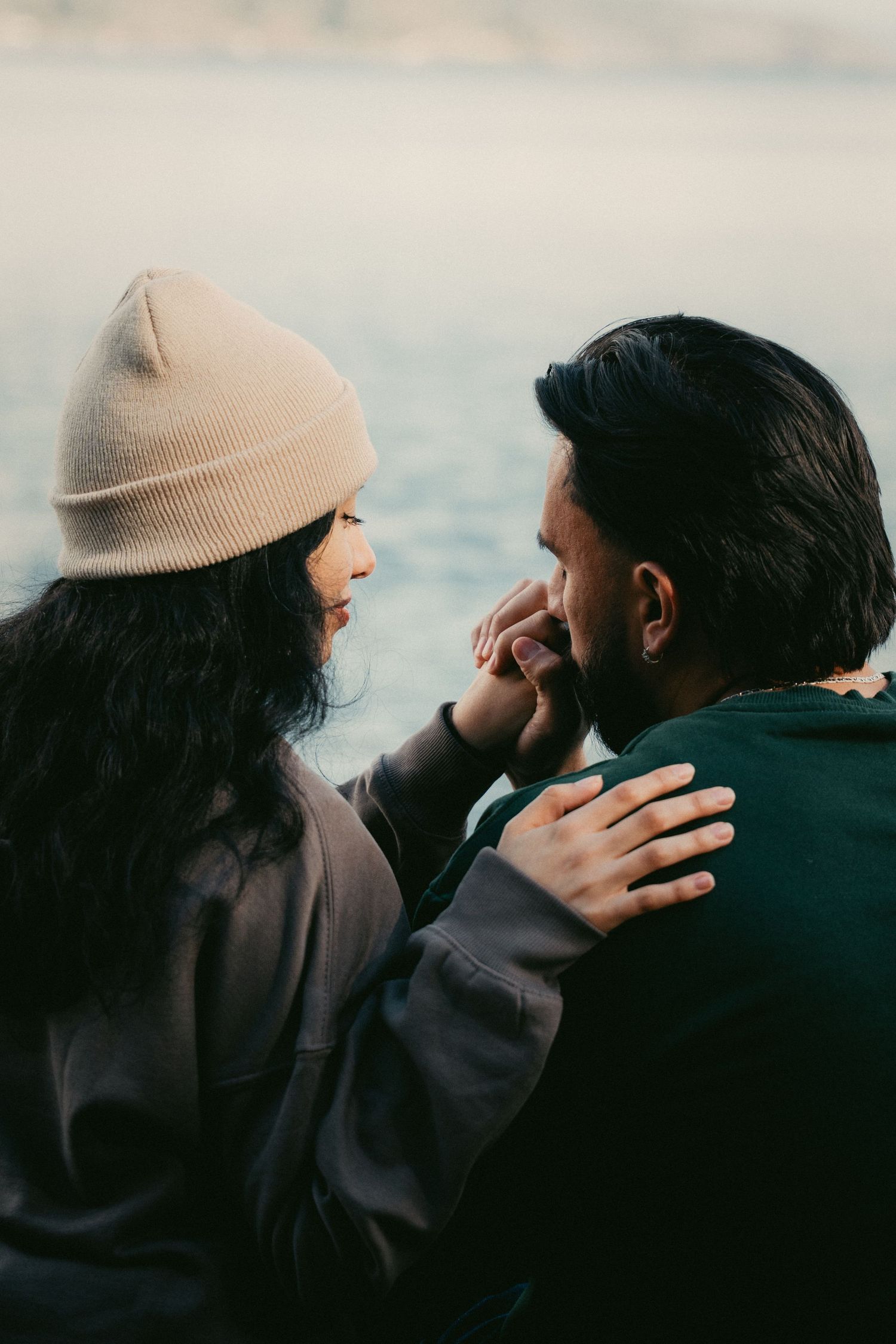A couple sits together near water, woman in beige beanie touching man's face while embracing him from behind.