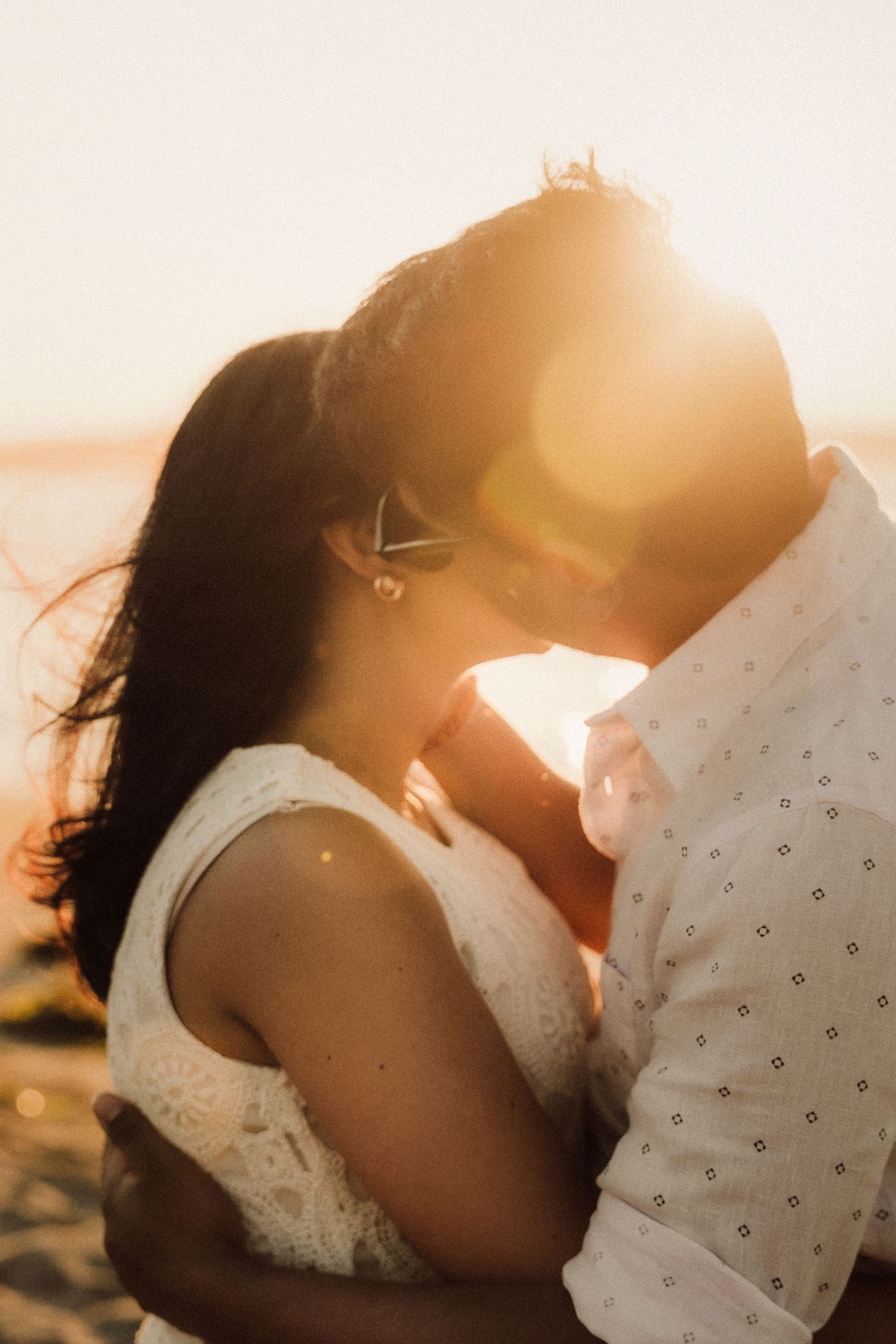 Couple sharing a romantic sunset kiss on the beach, bathed in warm golden light with wind-swept hair.