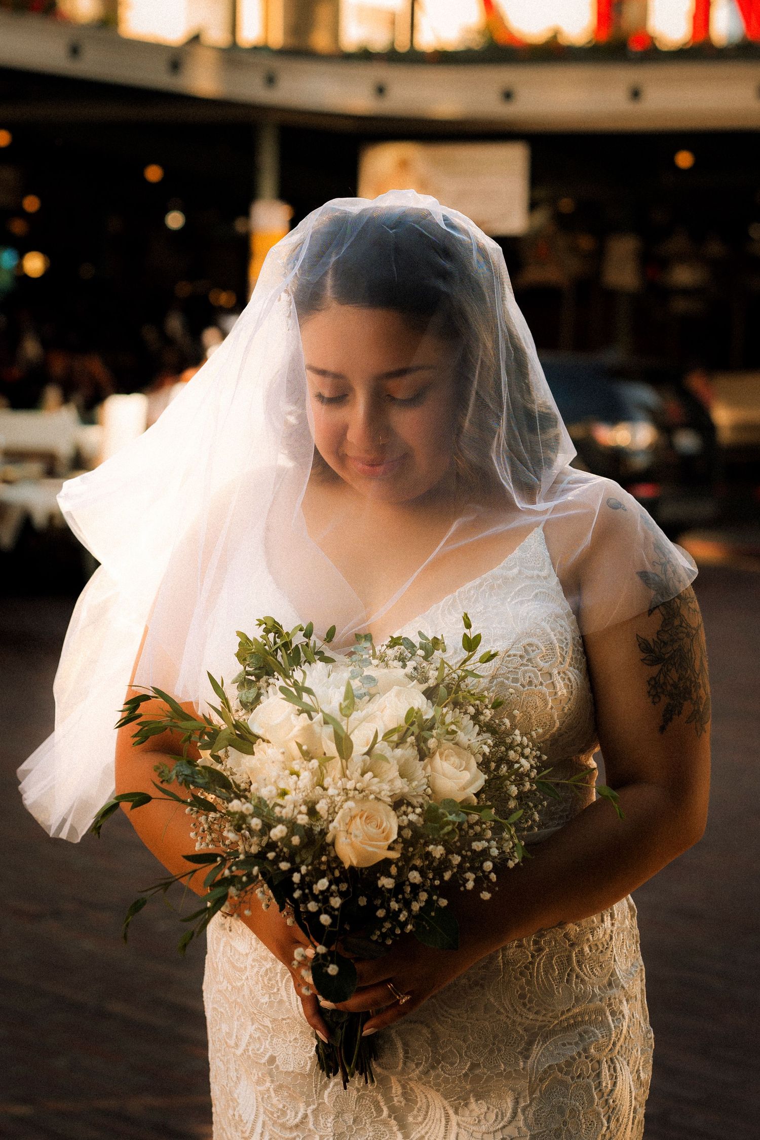 Bride in lace wedding dress and veil holds white rose bouquet, smiling softly outdoors at golden hour.