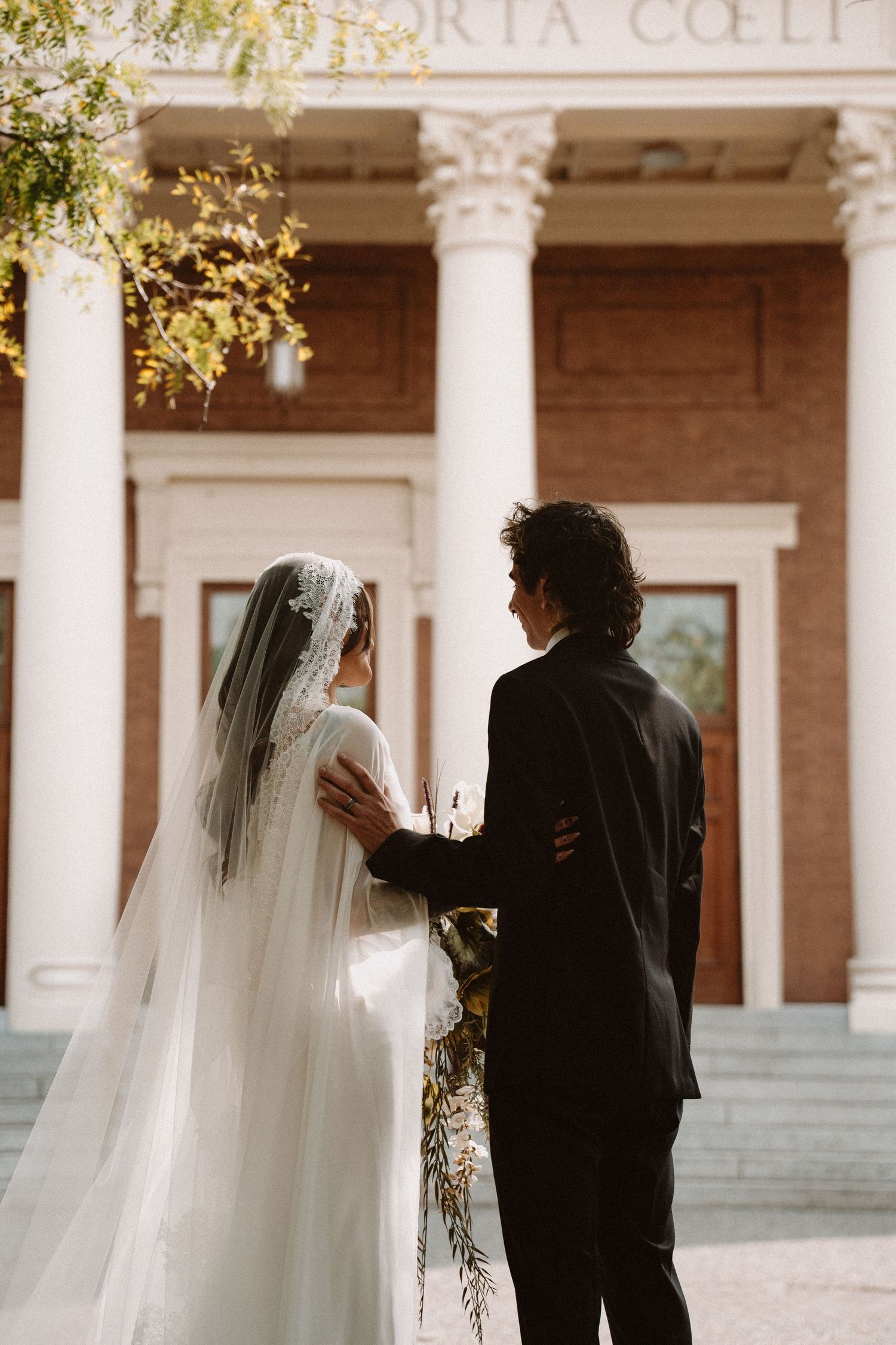 Bride in white gown and veil stands with groom in black suit before a grand church entrance on their wedding day.