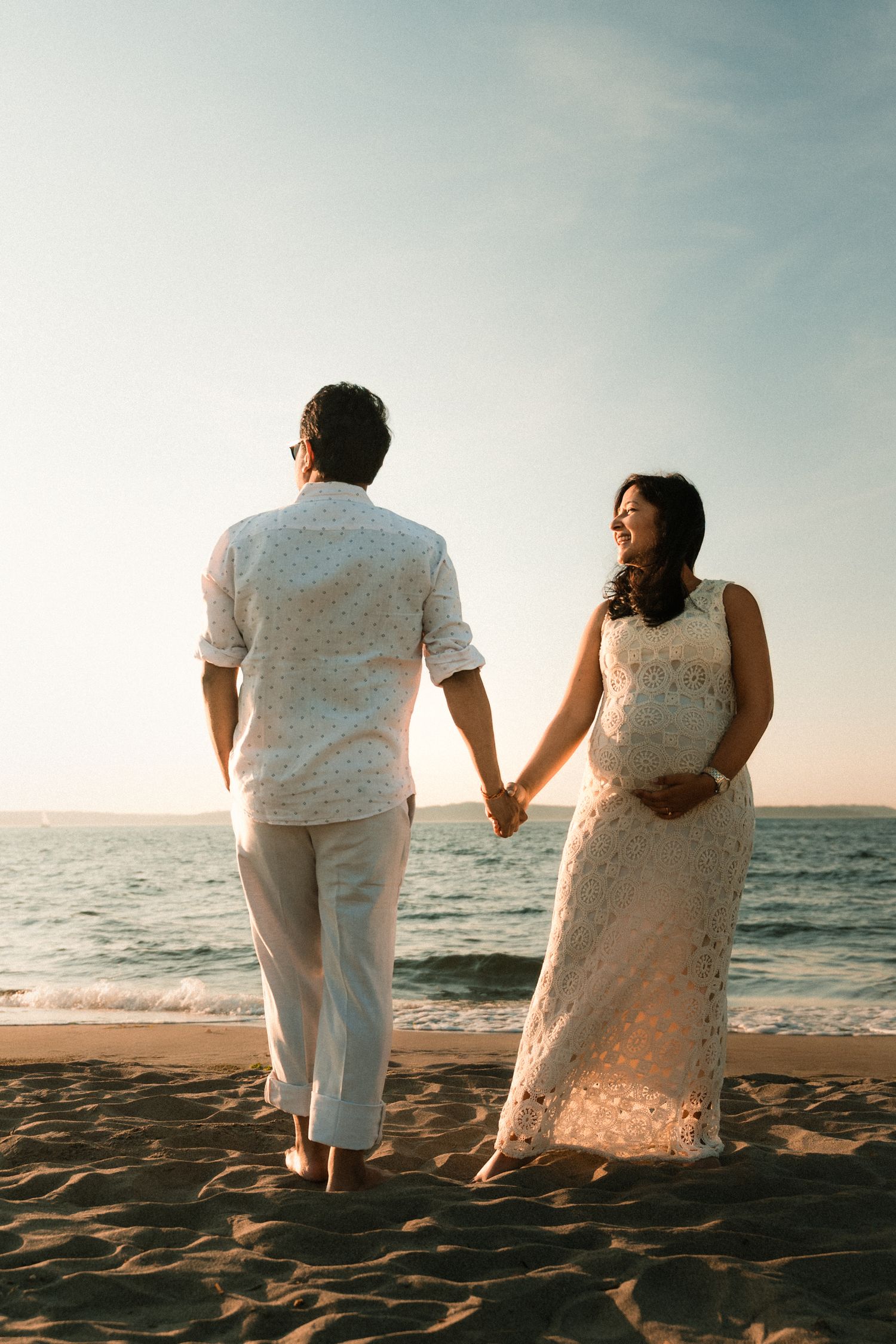 Couple holding hands on sandy beach at sunset, woman in white lace dress smiling, man in white shirt and pants.