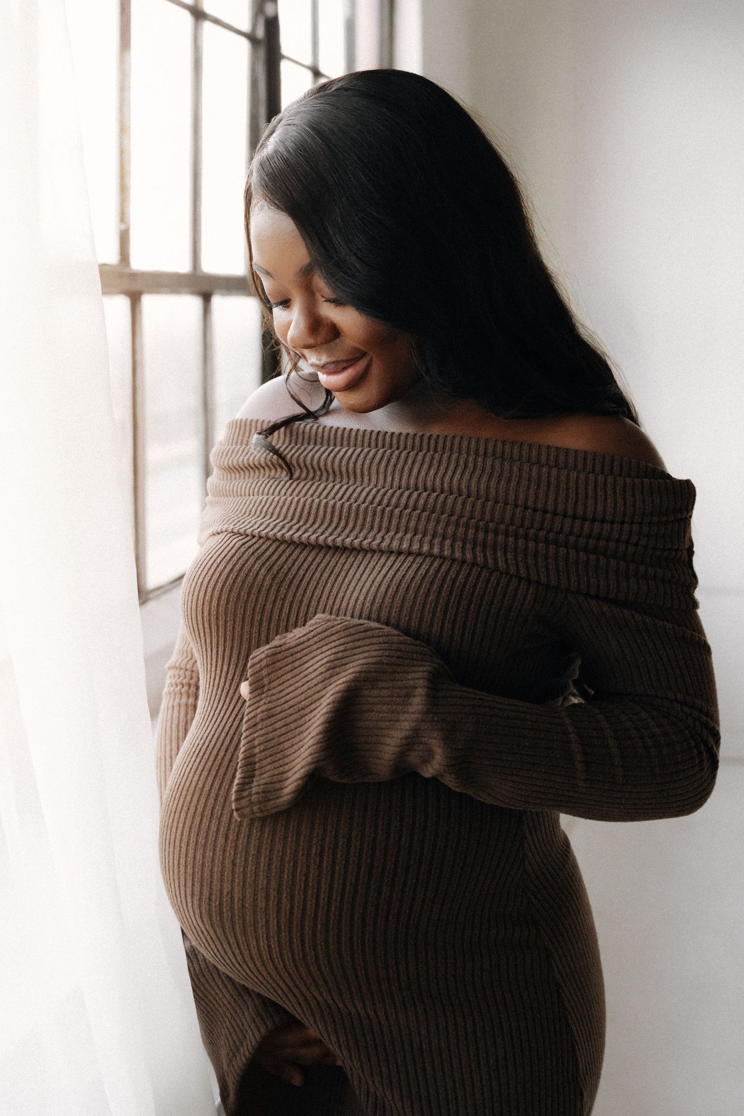 Smiling pregnant Black woman in brown off-shoulder ribbed dress cradling her baby bump by a sunlit window.