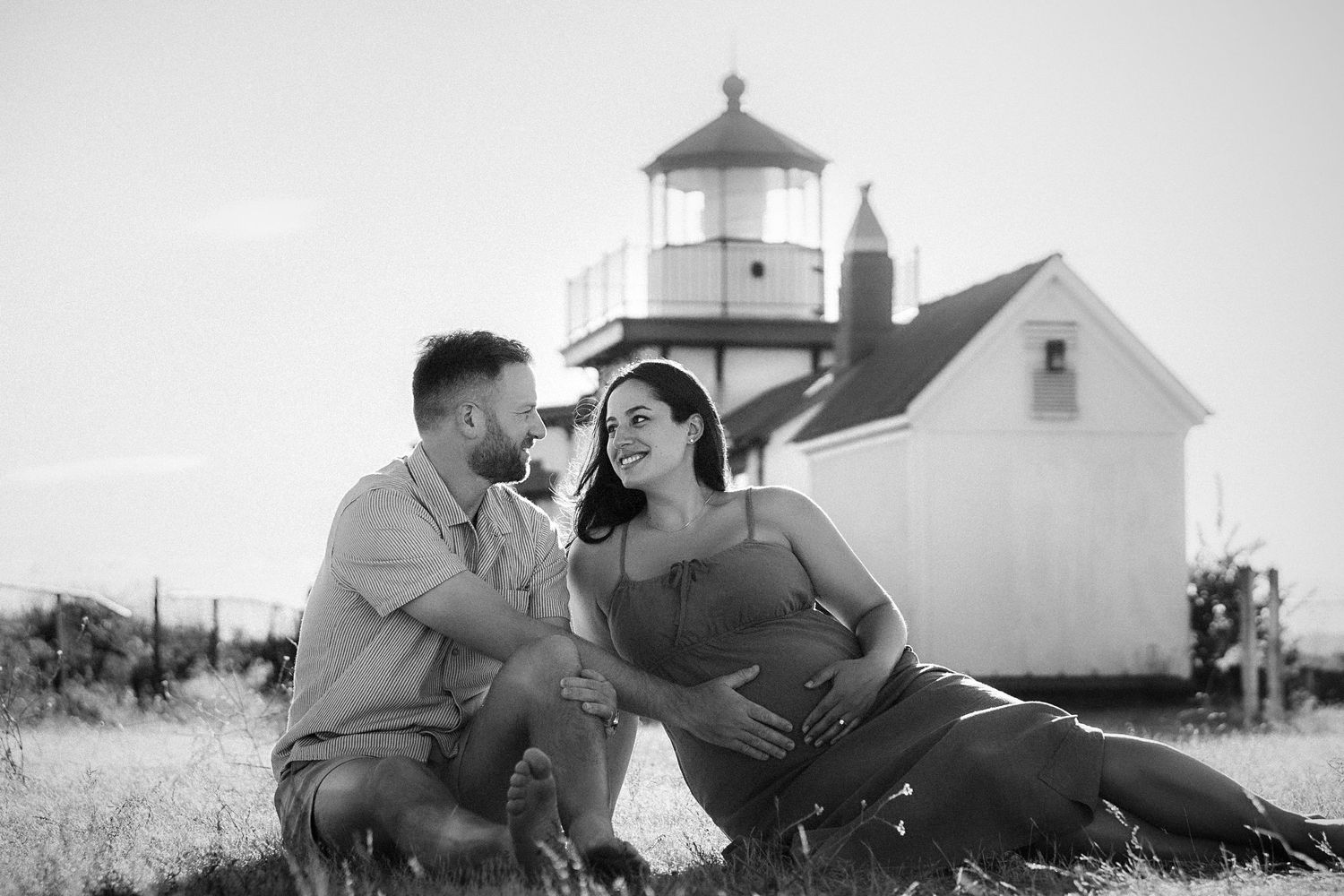 Black and white maternity photo of couple sitting in grass near a lighthouse, woman cradling baby bump.