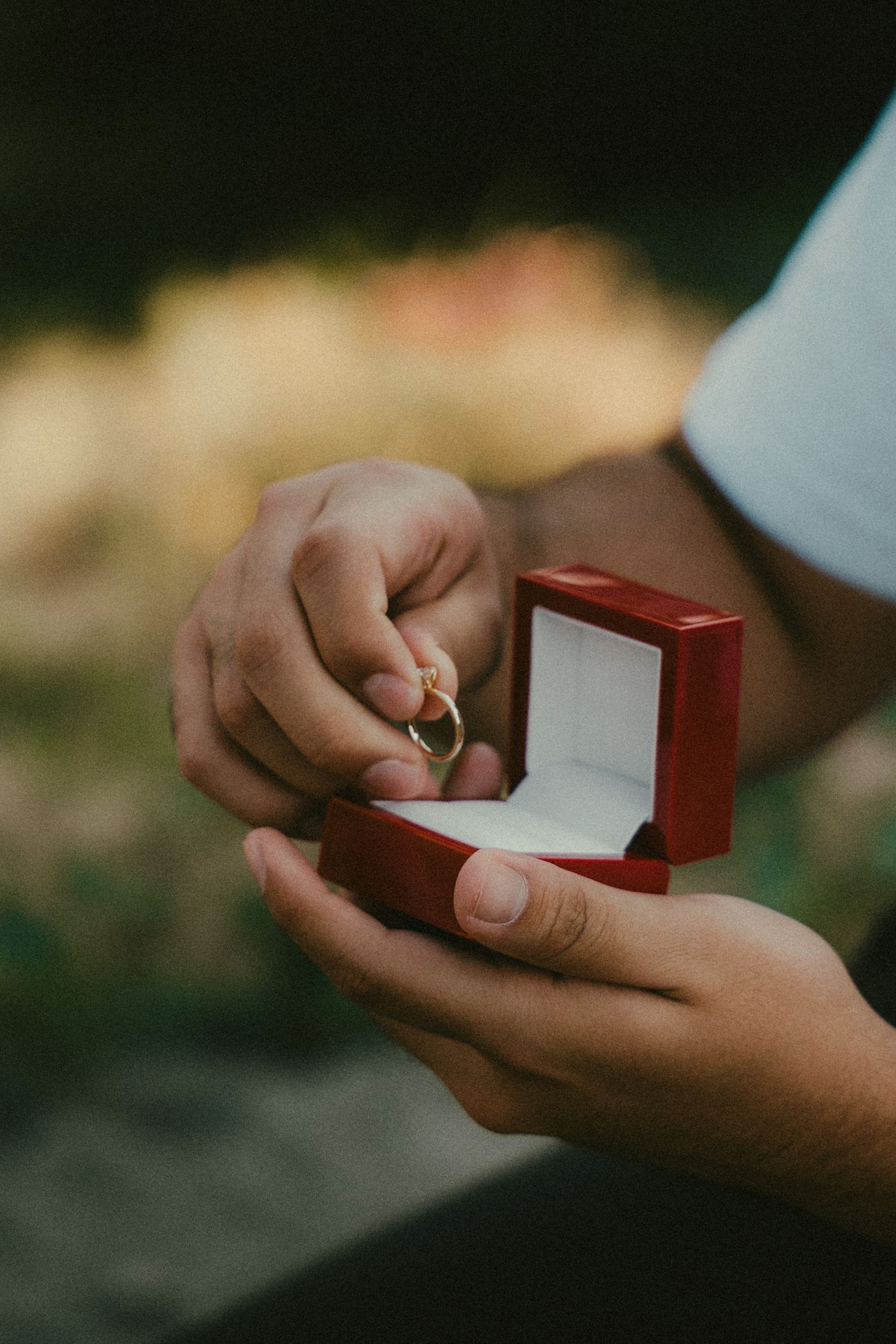 Person holding a red jewelry box with a gold engagement ring during an outdoor marriage proposal.