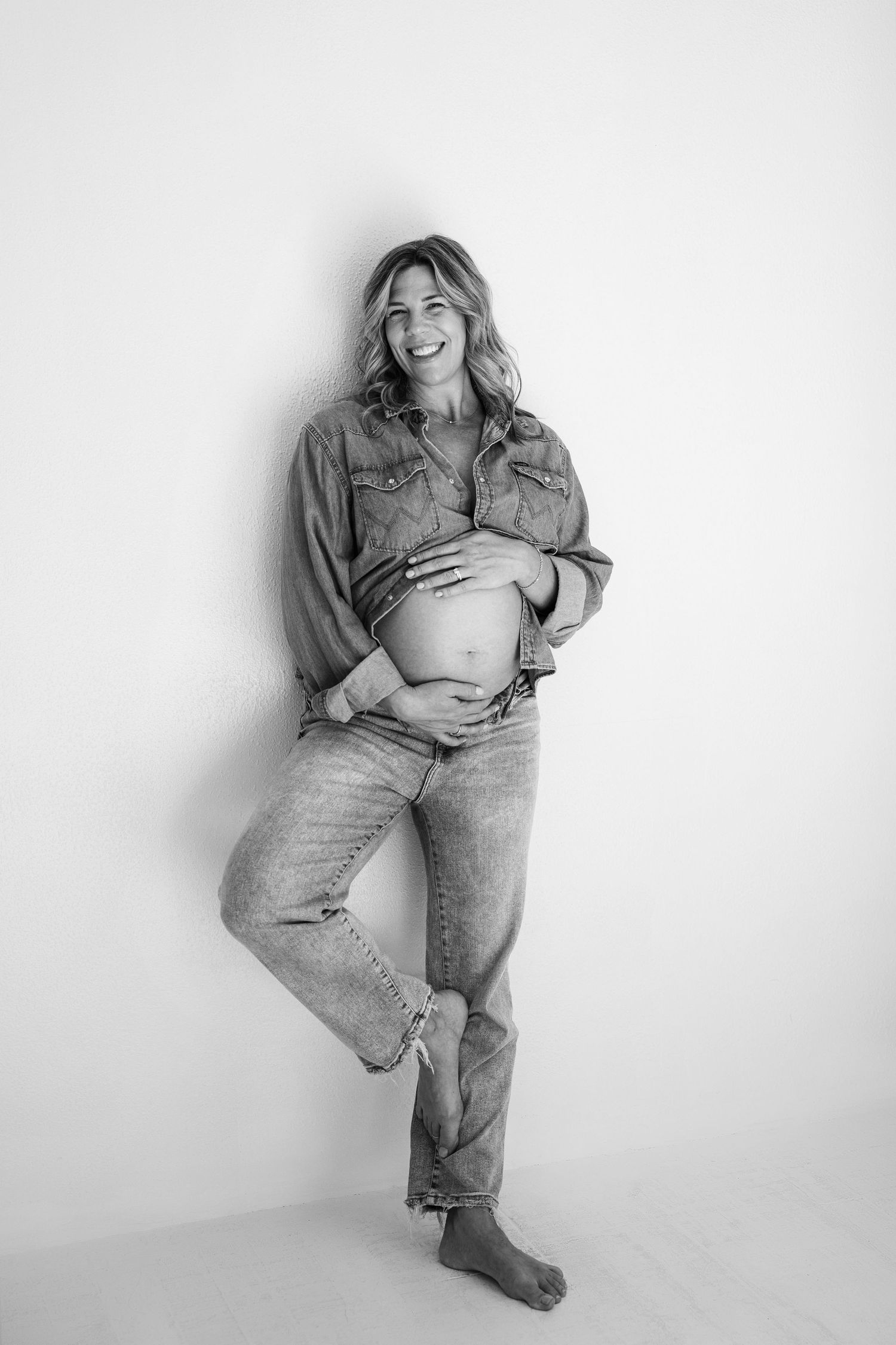 Black and white photo of smiling pregnant woman in denim shirt and jeans holding baby bump against white wall.