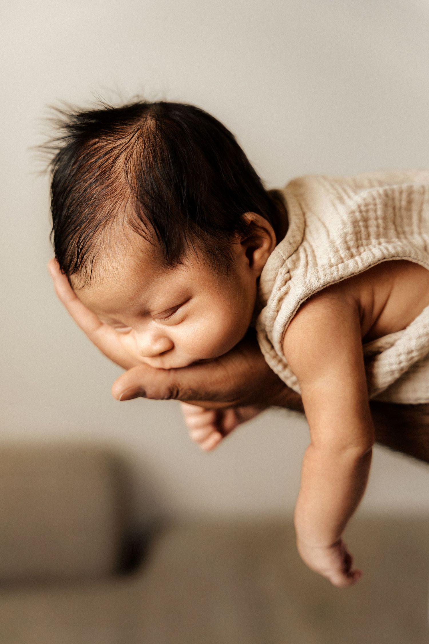 A newborn baby with dark hair sleeps peacefully while being cradled in an adult's hand, wearing a cream romper.