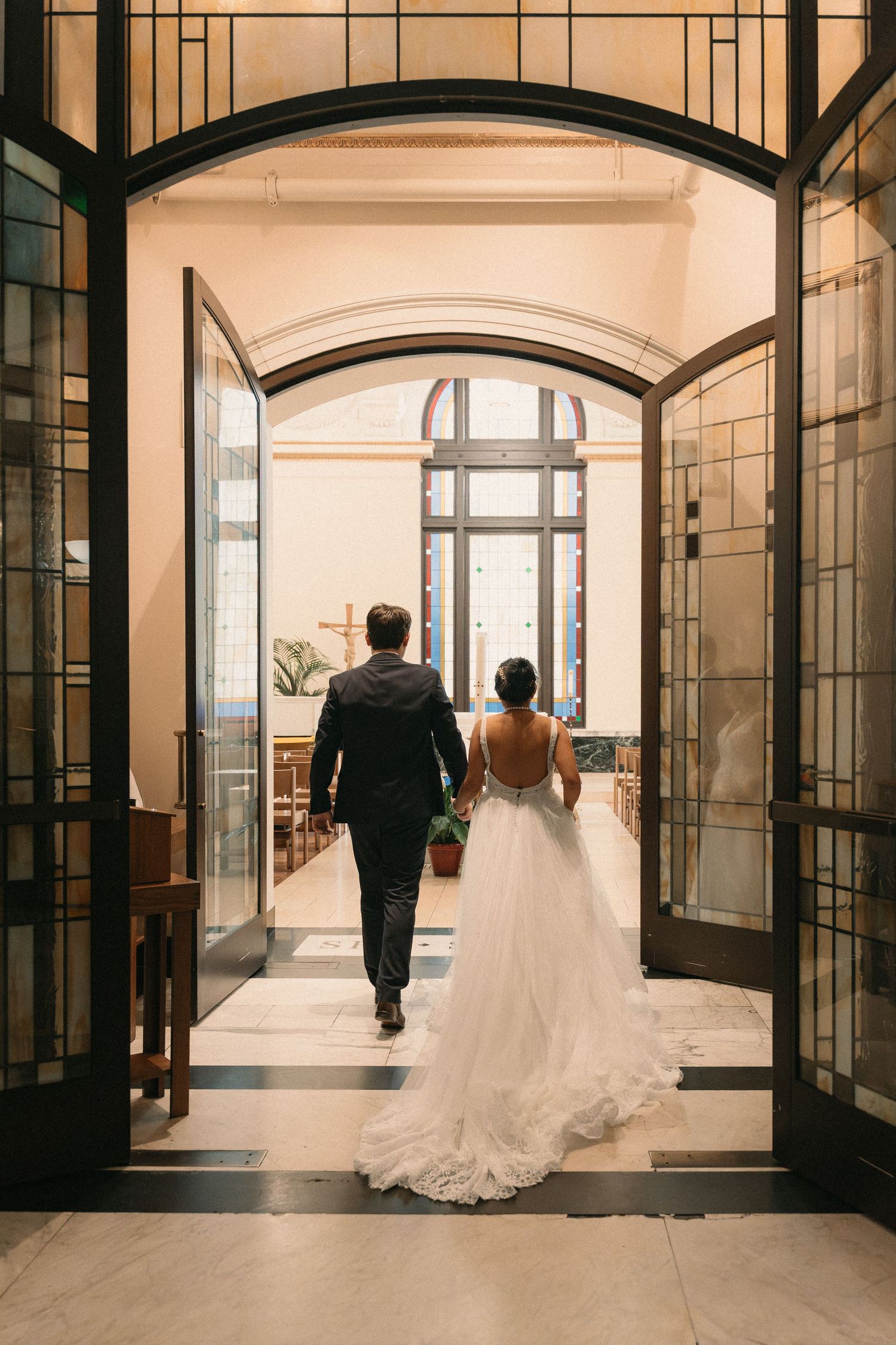 Bride and groom walking through elegant arched church doors, bride in backless white gown with flowing train.