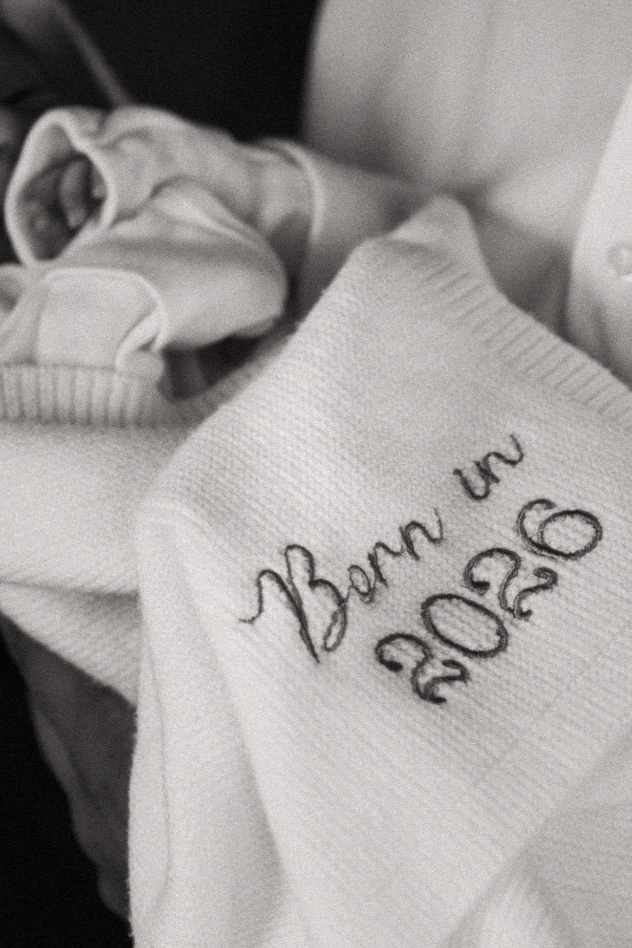 Black and white photo of a white knit baby blanket embroidered with Born in 2026 next to newborn baby clothes.