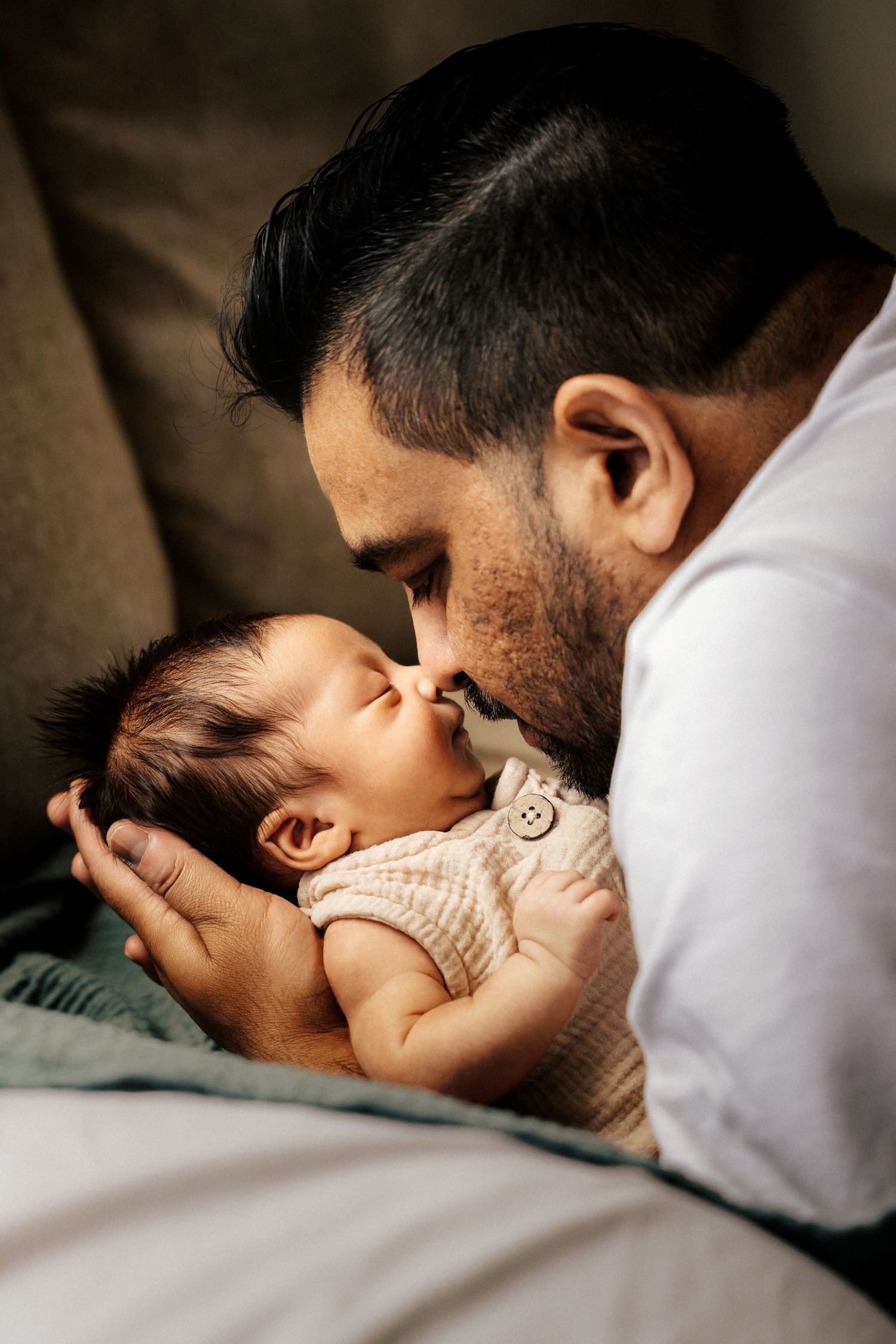 A bearded father tenderly kisses his sleeping newborn baby dressed in a cream knit onesie.