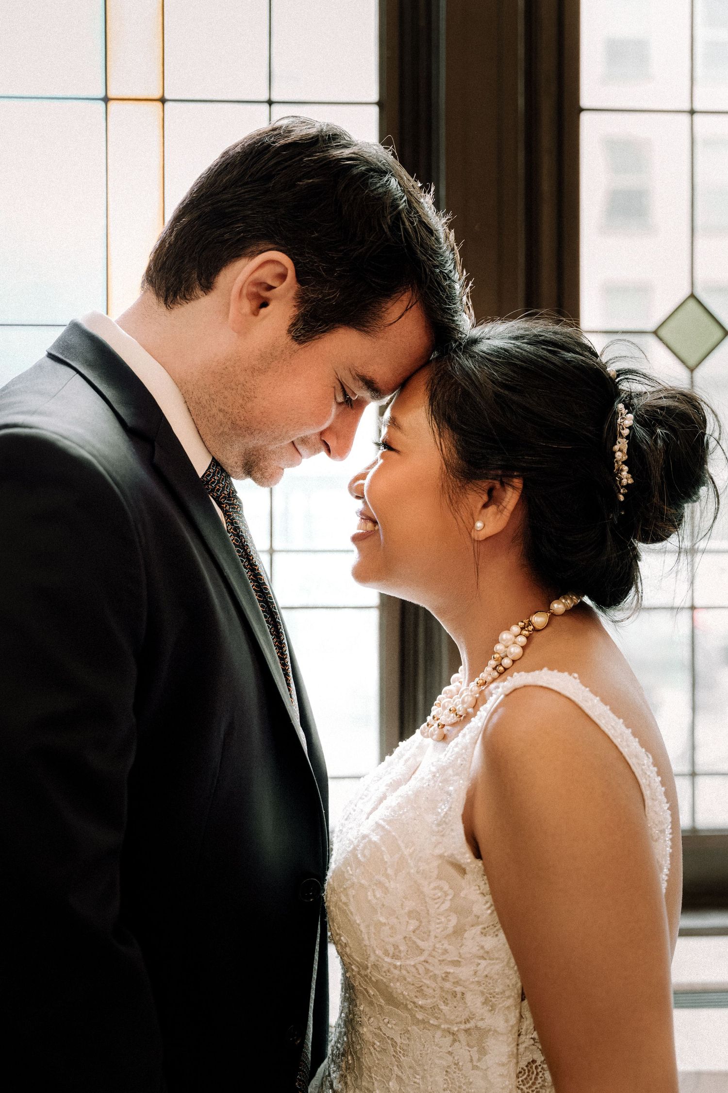 A bride and groom share an intimate forehead touch, smiling softly in wedding attire near a stained glass window.
