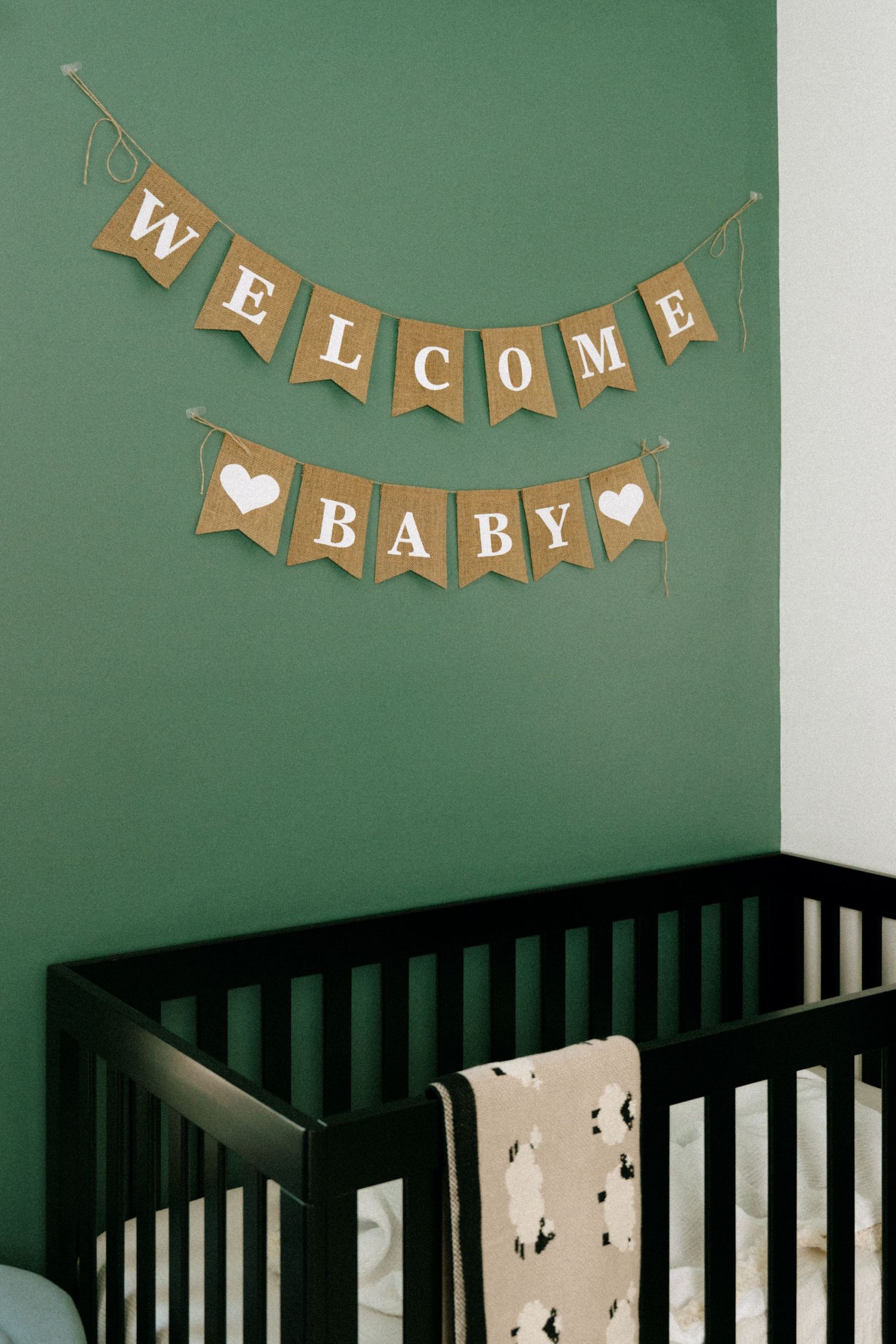 A dark wood baby crib beneath a burlap Welcome Baby banner on a green nursery wall.