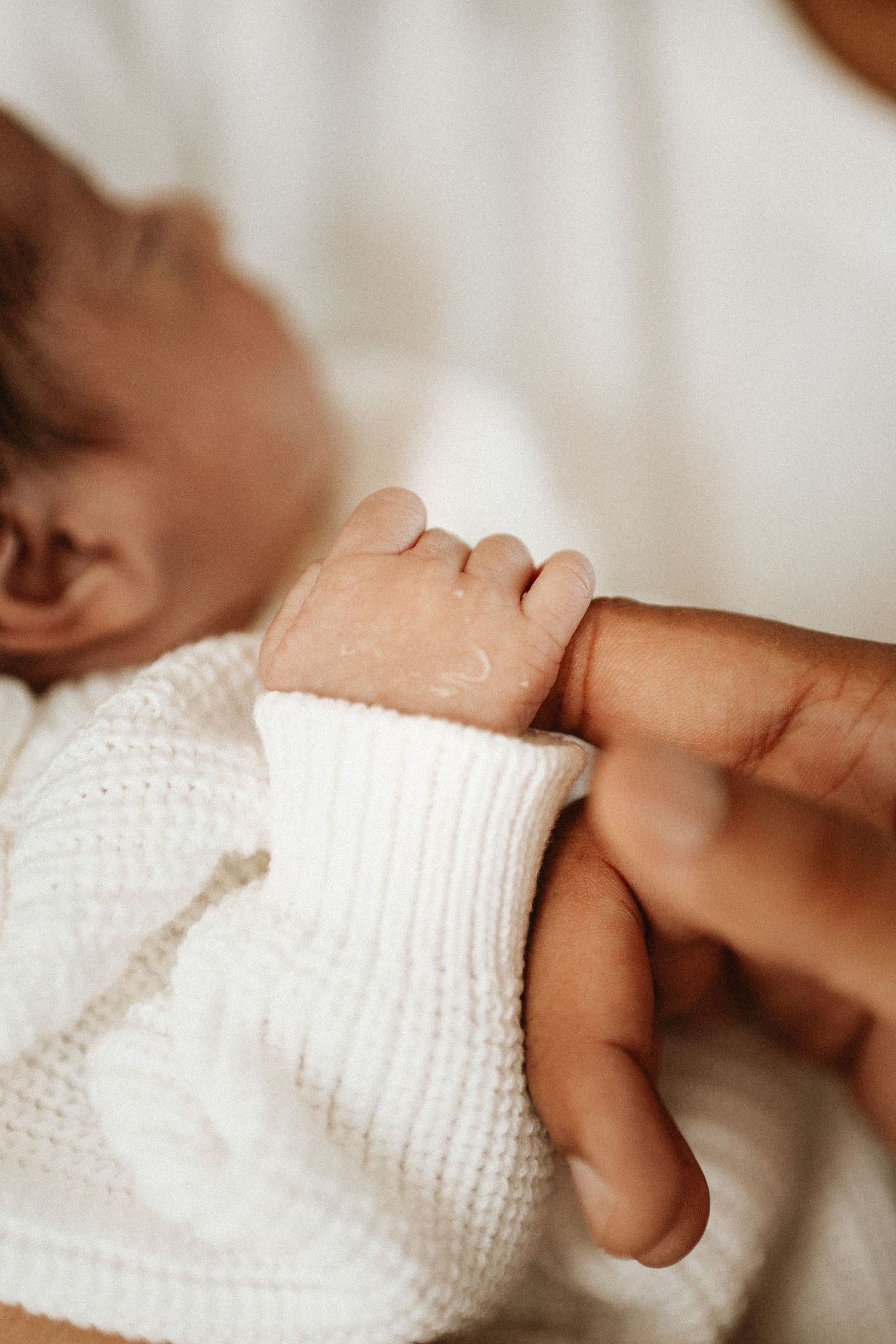 Newborn baby in white knit outfit grasping an adult's finger, close-up of tiny hand holding parent's thumb.