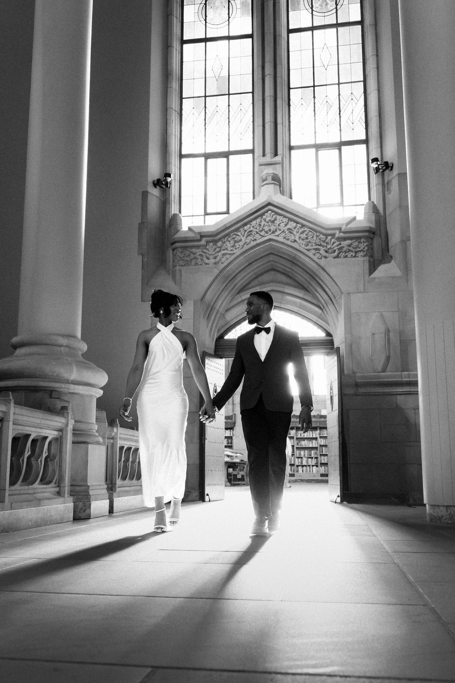 Black and white photo of a couple holding hands walking through an elegant church archway in wedding attire.