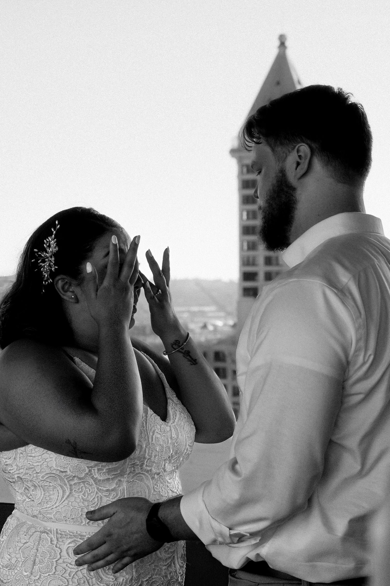 Black and white photo of a bride covering her face emotionally as groom stands before her on a rooftop wedding day.