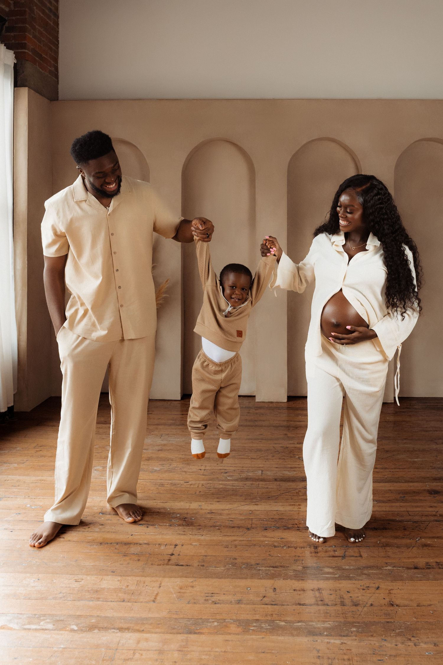 A joyful Black family in matching beige outfits lifting their toddler while the mother shows her pregnant belly.