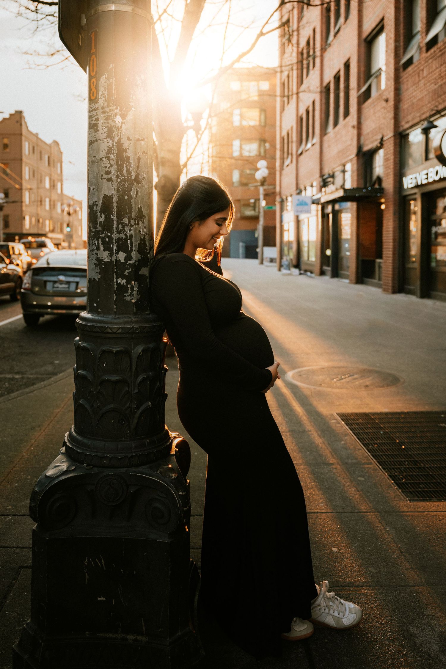 Pregnant woman in black dress leaning on street lamp post at golden hour in urban city setting.