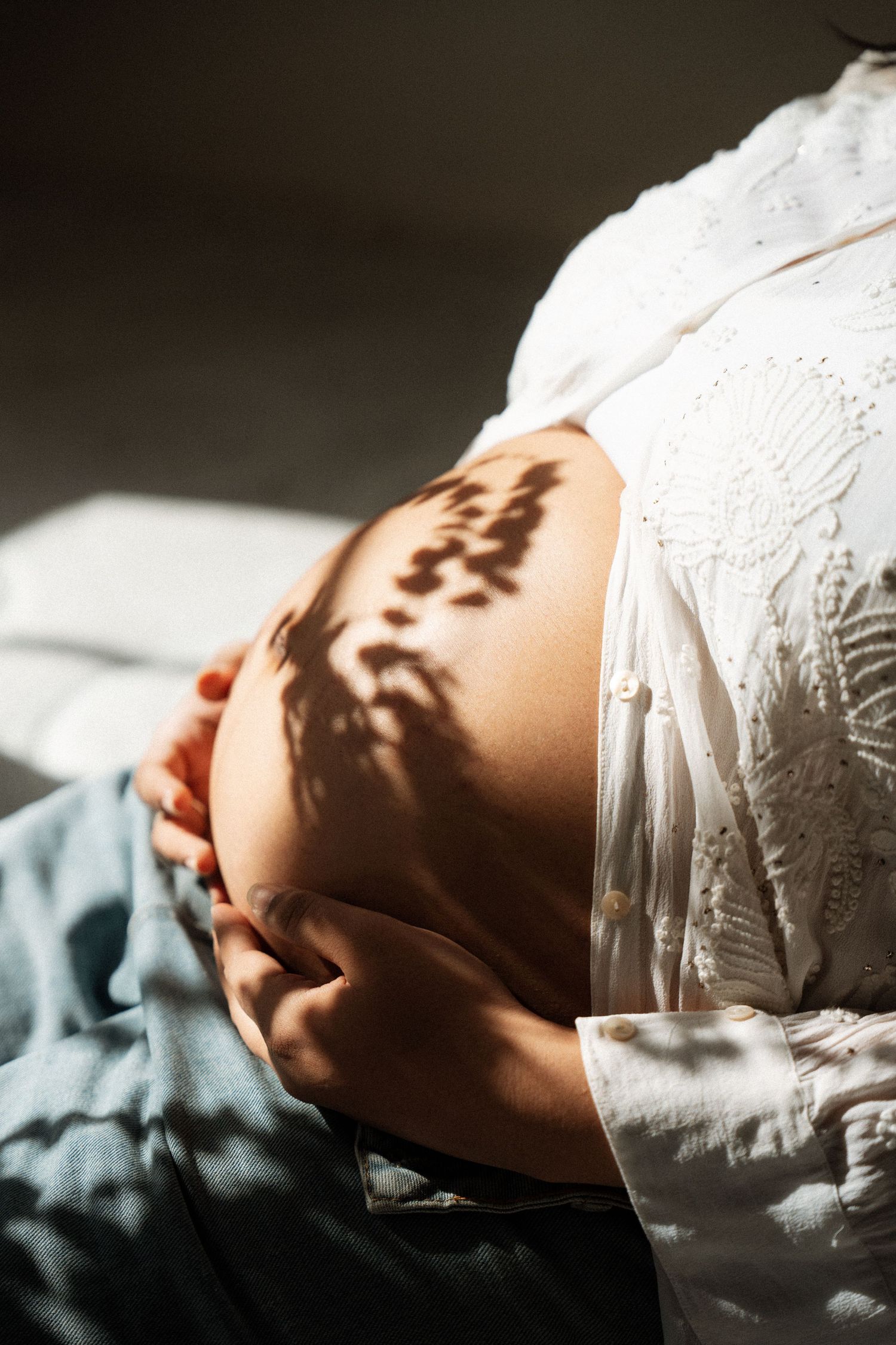 Pregnant woman in white lace top cradling bare baby bump with dappled sunlight shadows in serene maternity photo.