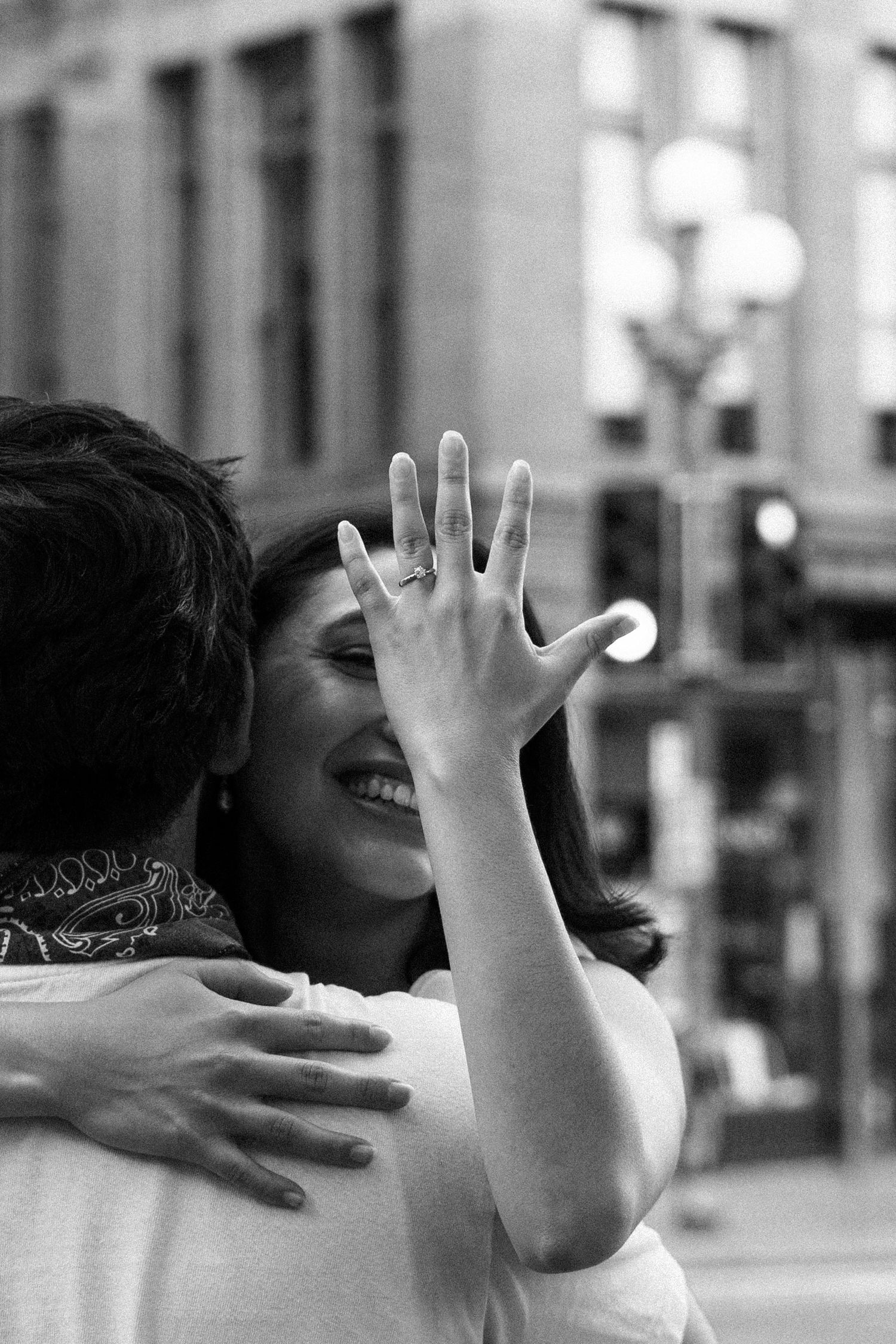 Black and white photo of a smiling woman hugging someone while showing off her engagement ring to the camera.